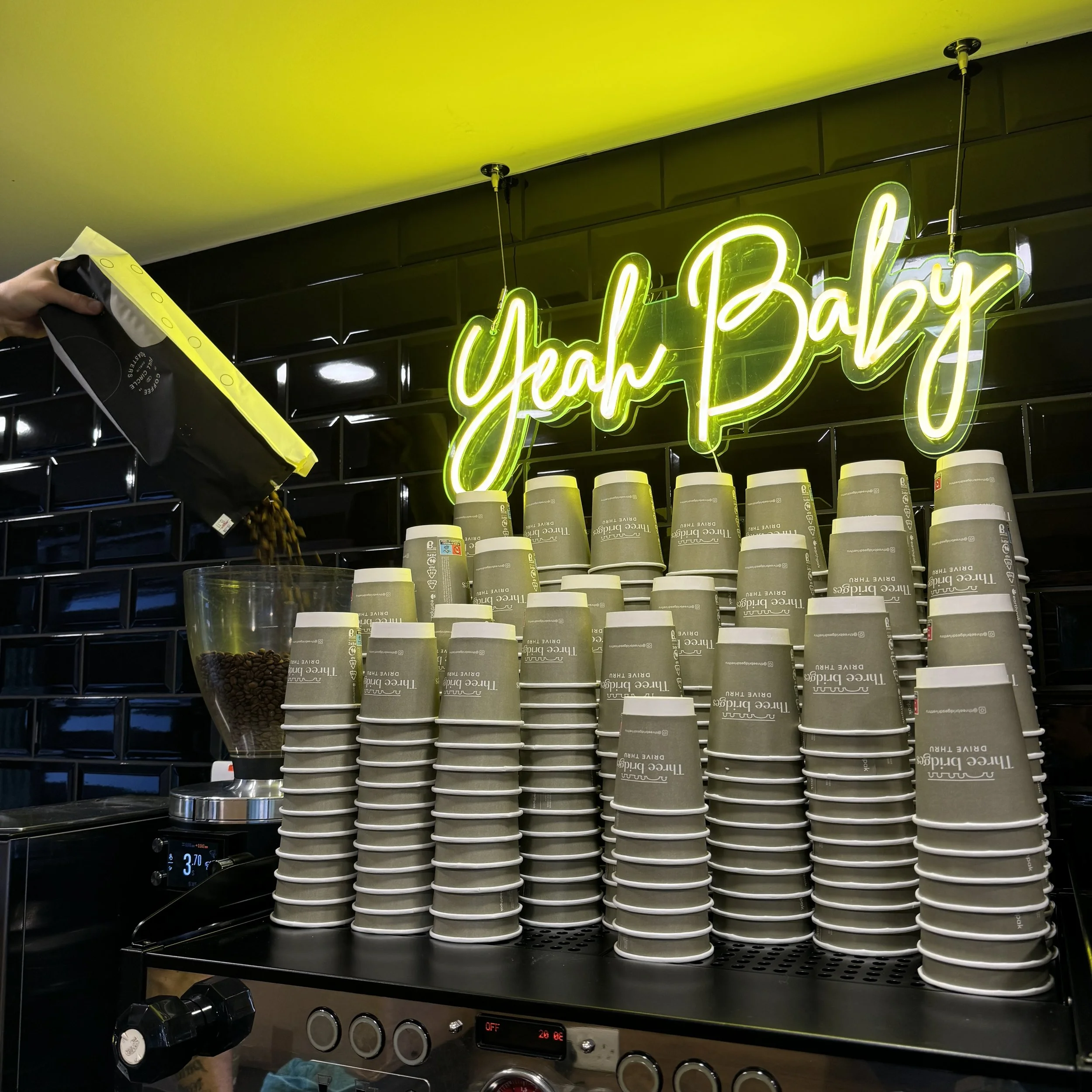 Stacked coffee cups beneath a neon sign that reads 'Yah Baby' in a coffee shop.