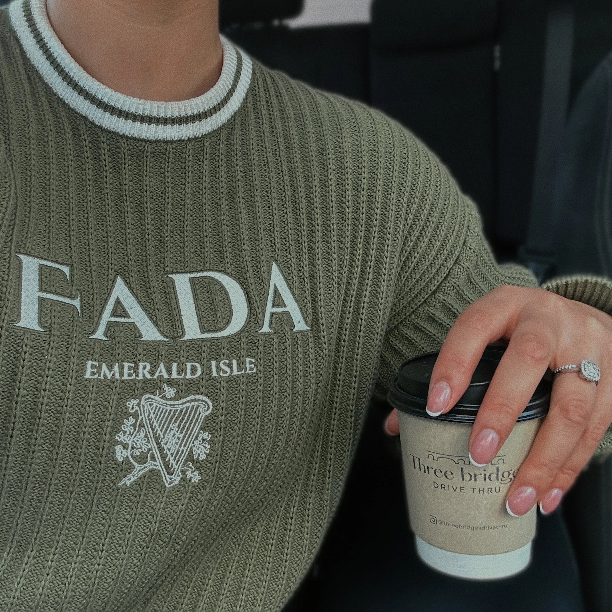 Person wearing green sweater with 'FADA Emerald Isle' text holding a coffee cup from Three Bridges Drive Thru, showing rings on finger.