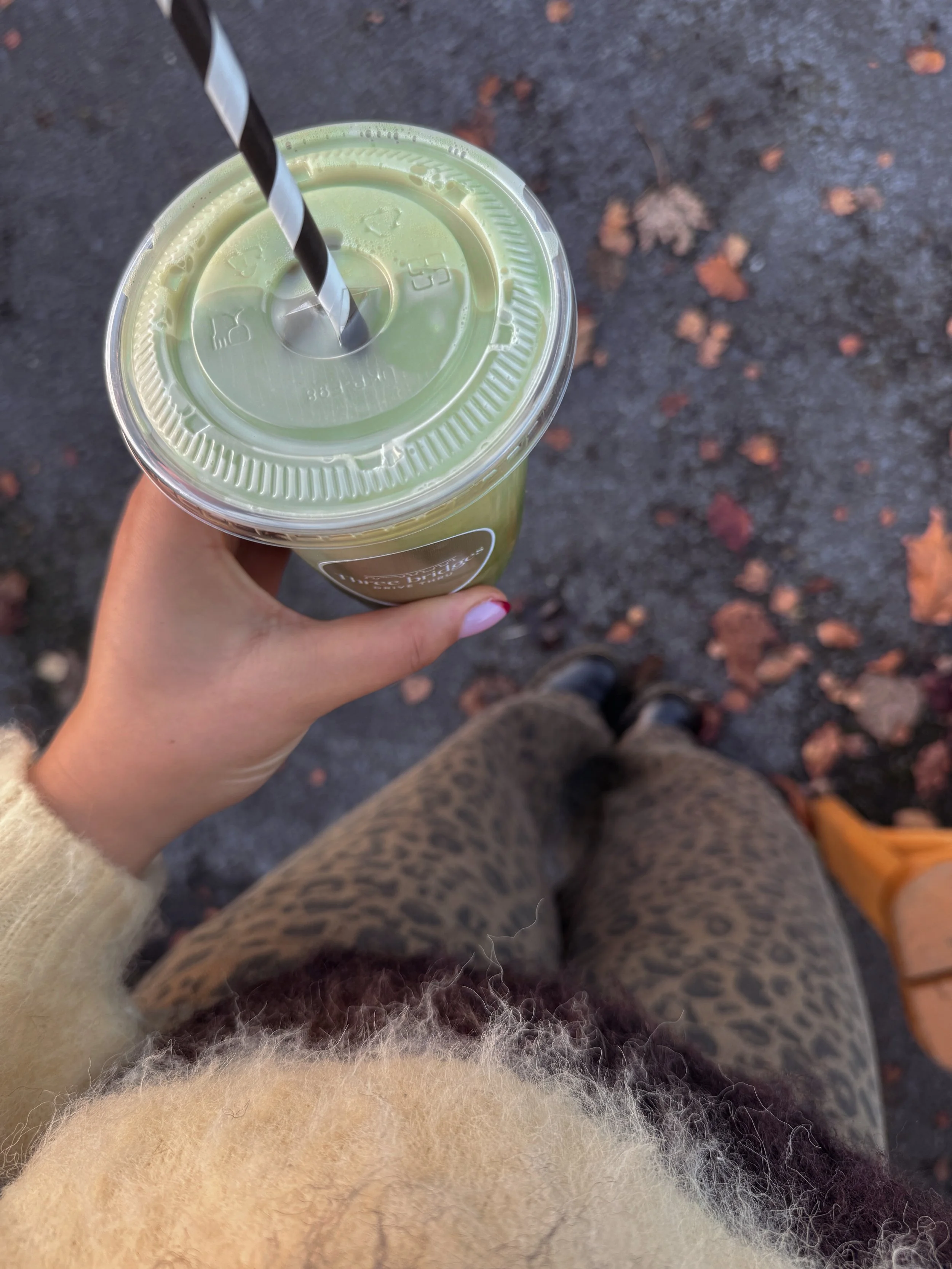 A person holding a plastic cup with a green smoothie and a black and white striped straw, seen from above on a pavement with fallen leaves.