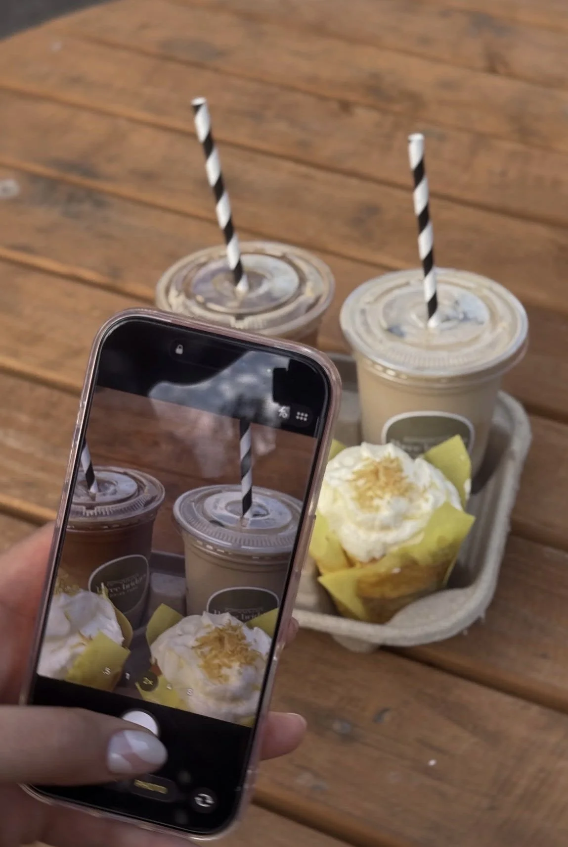 Person taking a photo of two iced coffee drinks and a dessert with a smartphone. The drinks have black-and-white striped straws, and the dessert is topped with whipped cream and pineapple chunks. The tray with the items is on a wooden table.