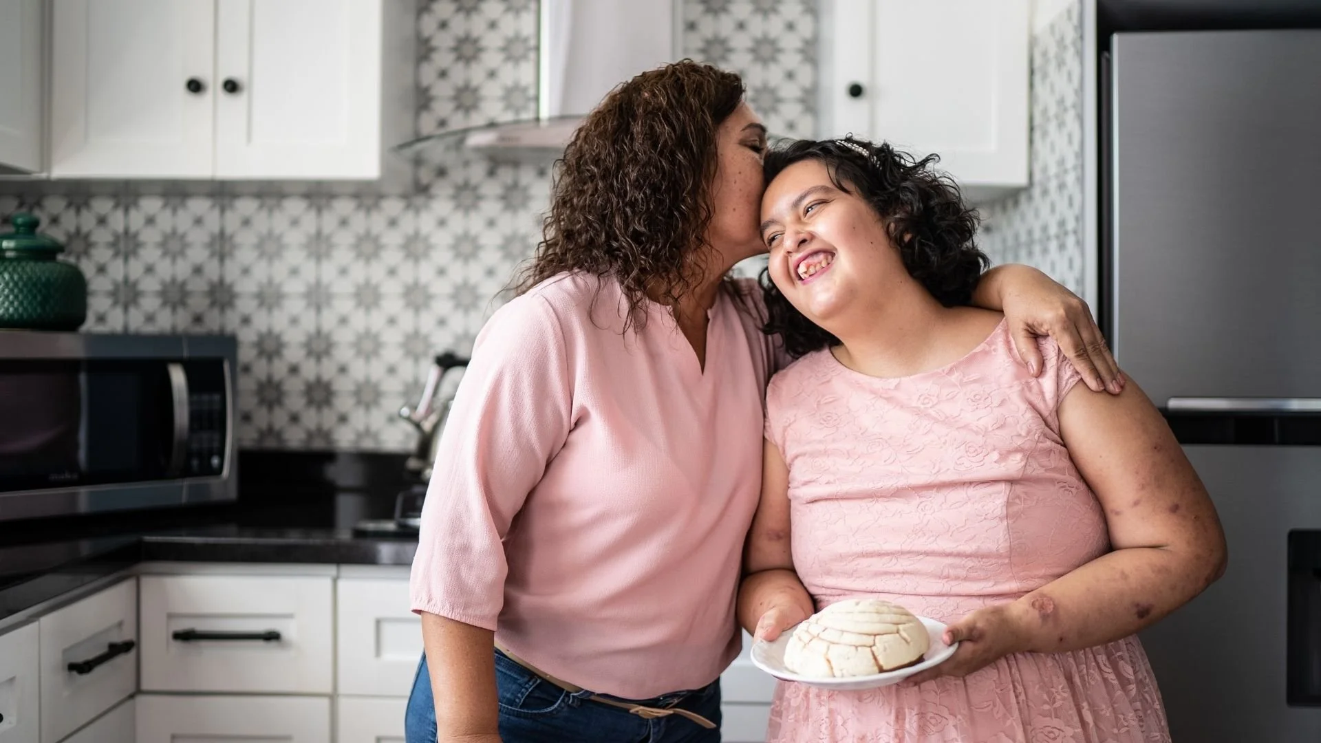 A woman and a girl sharing a moment in the kitchen, with the woman kissing the girl on the cheek as they both smile. The girl holds a plate with a dome-shaped cake, and the girl has visible scars and bruises on her arms.