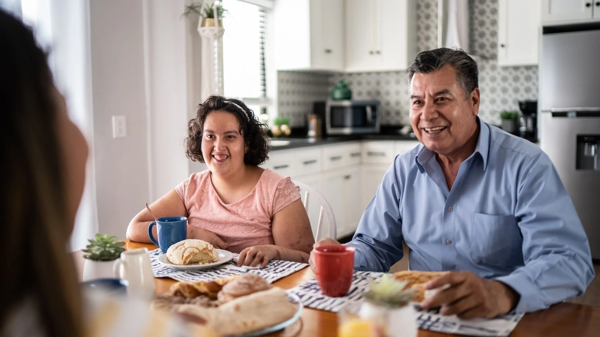 A family gathered around a kitchen table enjoying breakfast with bread, coffee, and pastries, smiling and talking.