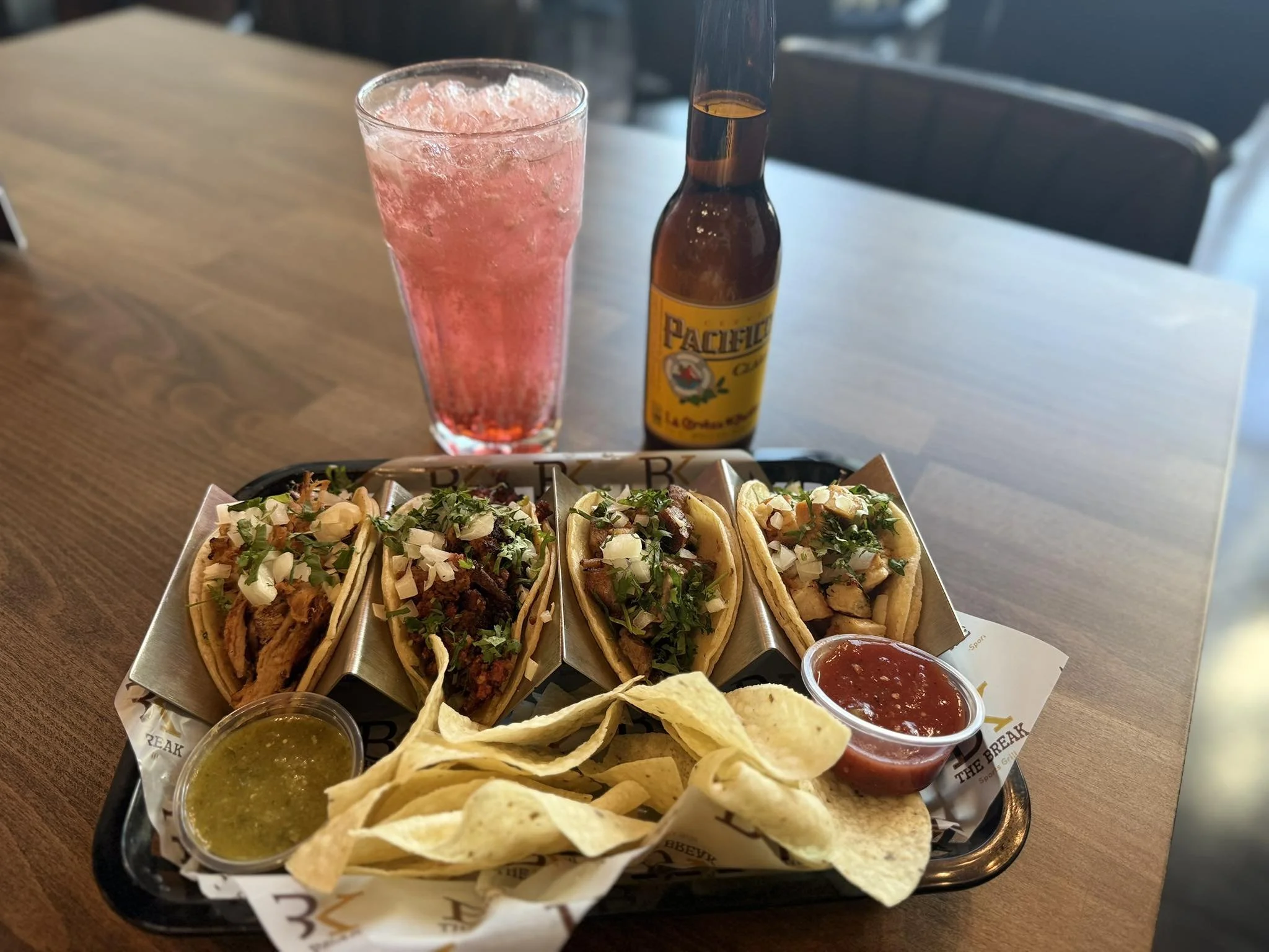 A tray with three tacos topped with chopped onions, cilantro, and meat, accompanied by potato chips, two small cups of salsa, and a green sauce. In the background, a pink drink and a bottle of Pacifico beer placed on a wooden table.