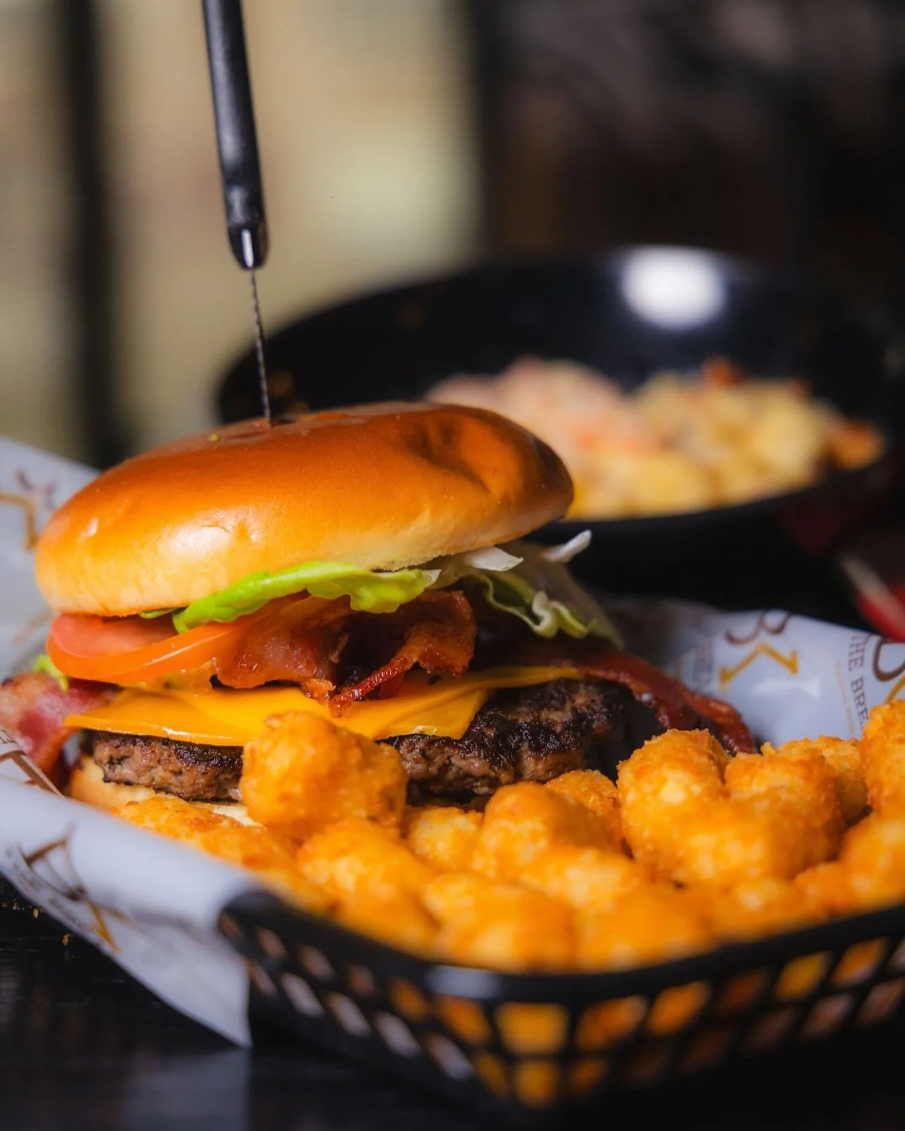 A cheeseburger with lettuce, tomato, bacon, and cheese, served with cheese puffs in a basket, with a black skillet in the background.