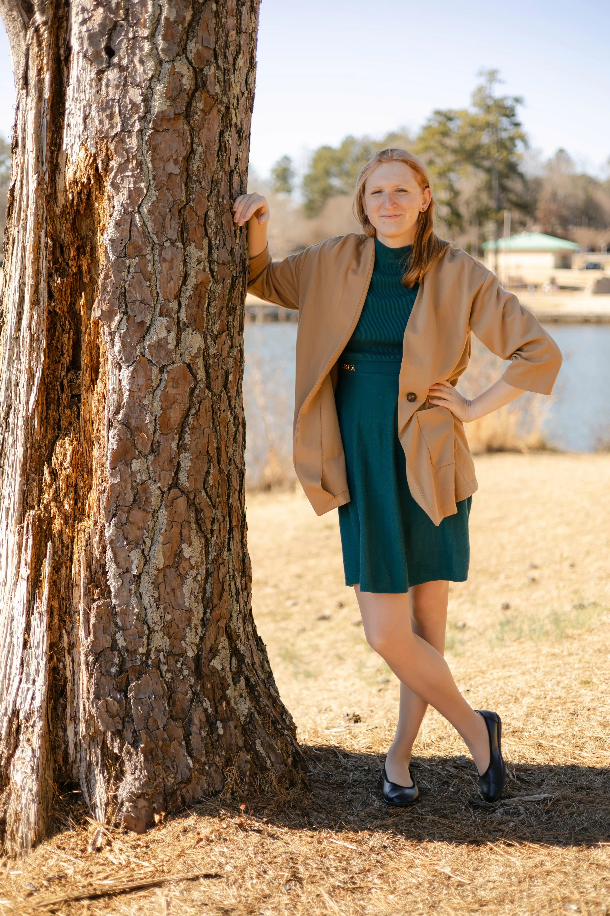 A woman in a green dress and tan blazer leans against a pine tree with her legs crossed. A lake and more trees are seen in the background.