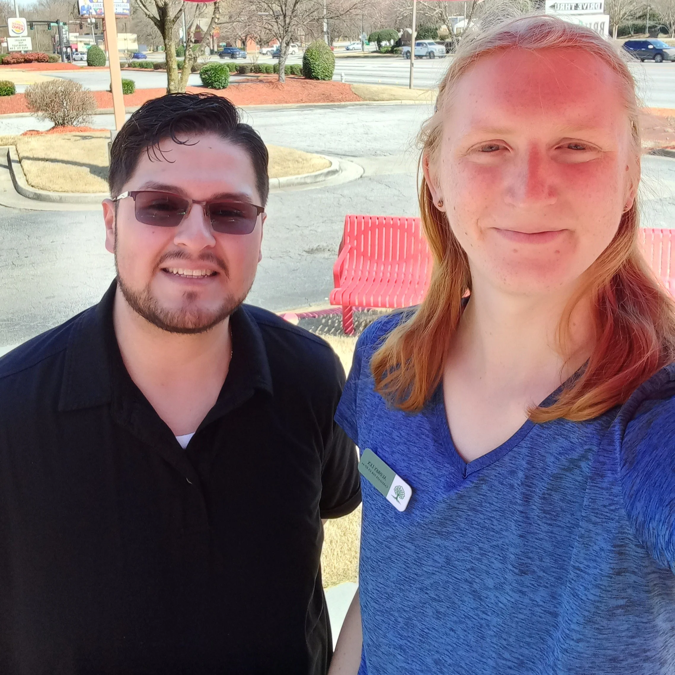 A man with short black hair, a goatee, glasses, and a black polo shirt stands next to a taller woman with long red hair in a blue T-shirt.