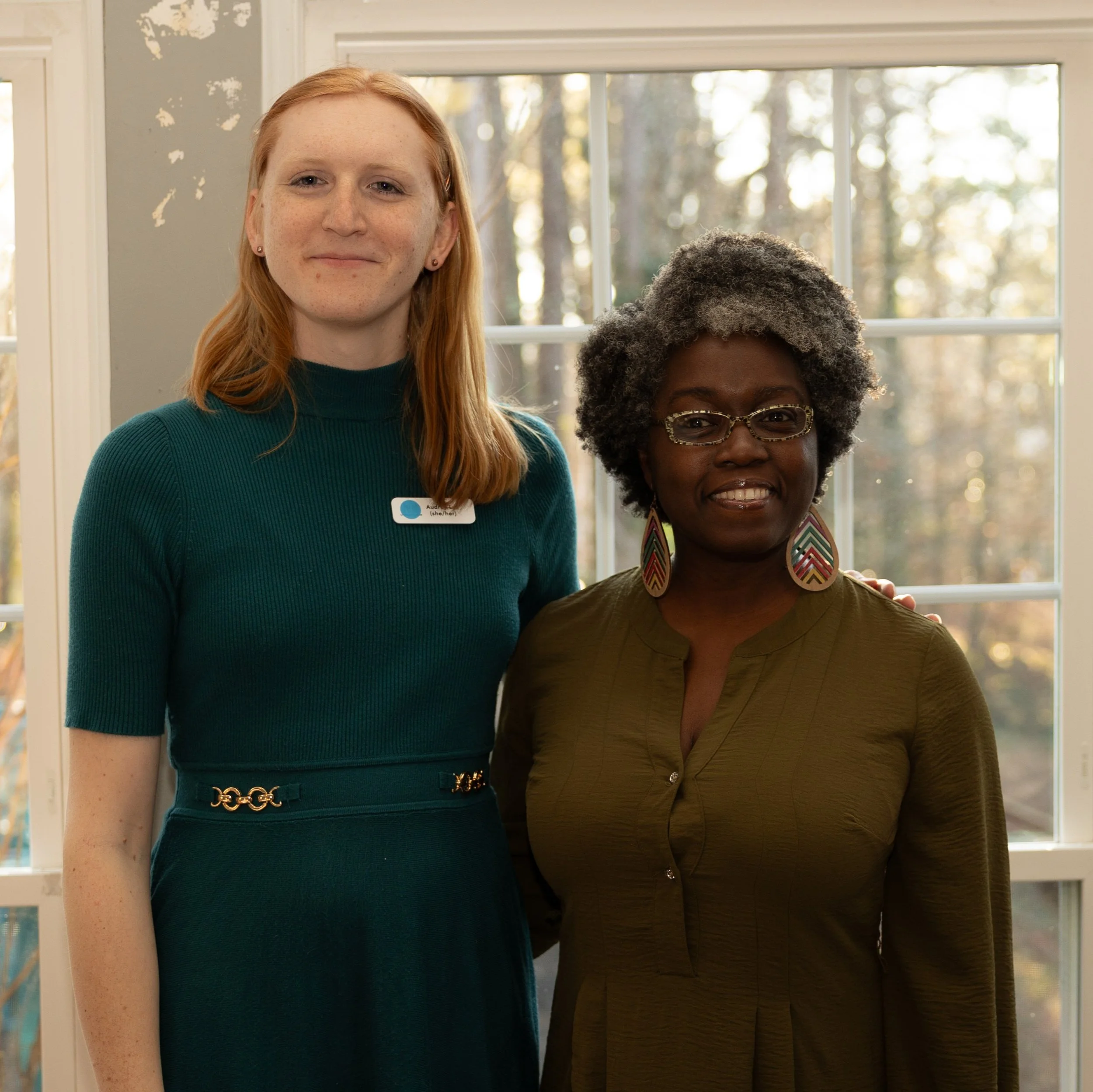 Two women smiling standing next to each other. The woman on the left is taller with long red hair and a bright green dress. The woman on the right is shorter with black hari, an olive colored dress, and large hoop earings.