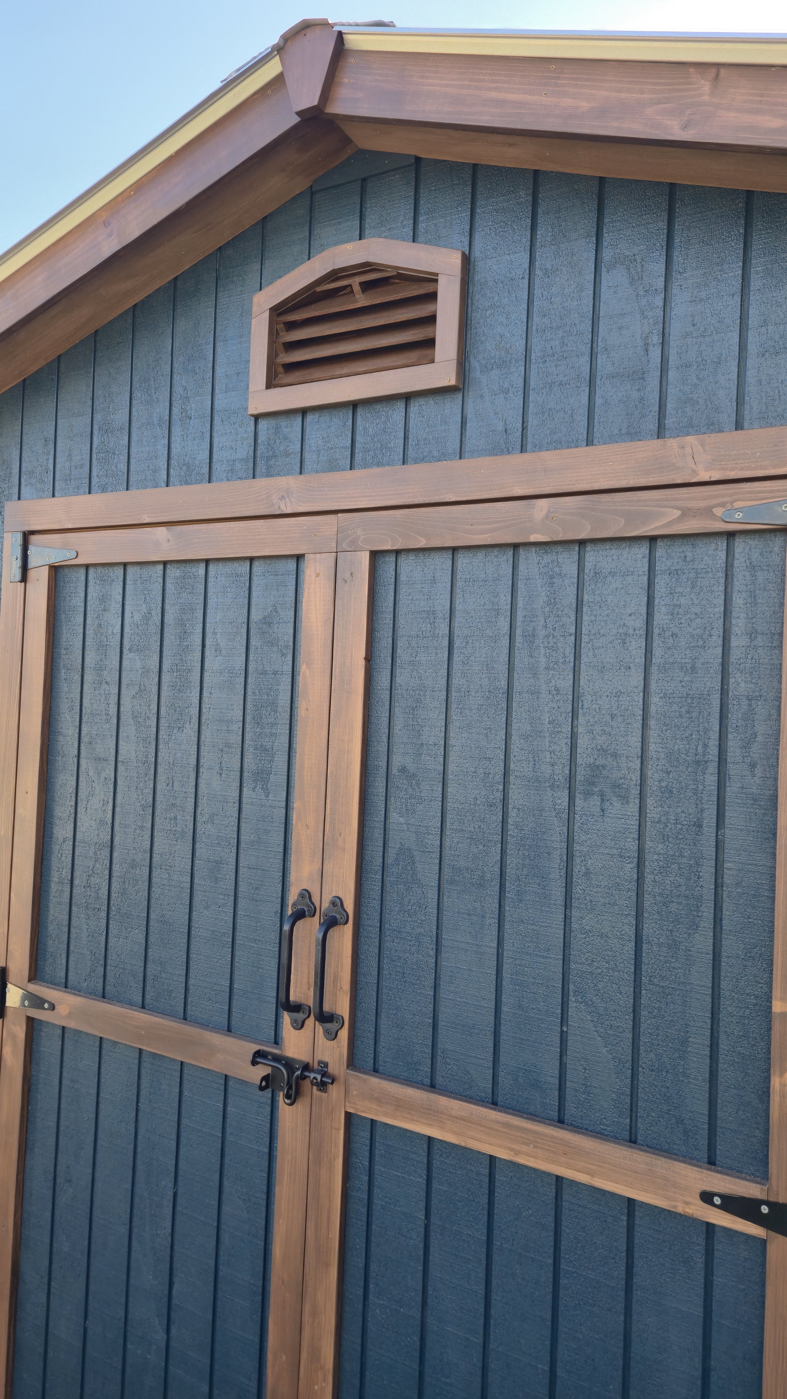 Detail photo of shed with wooden trim and black handles on double door, custom made gable roof ventilation opening with wooden slats above doors.