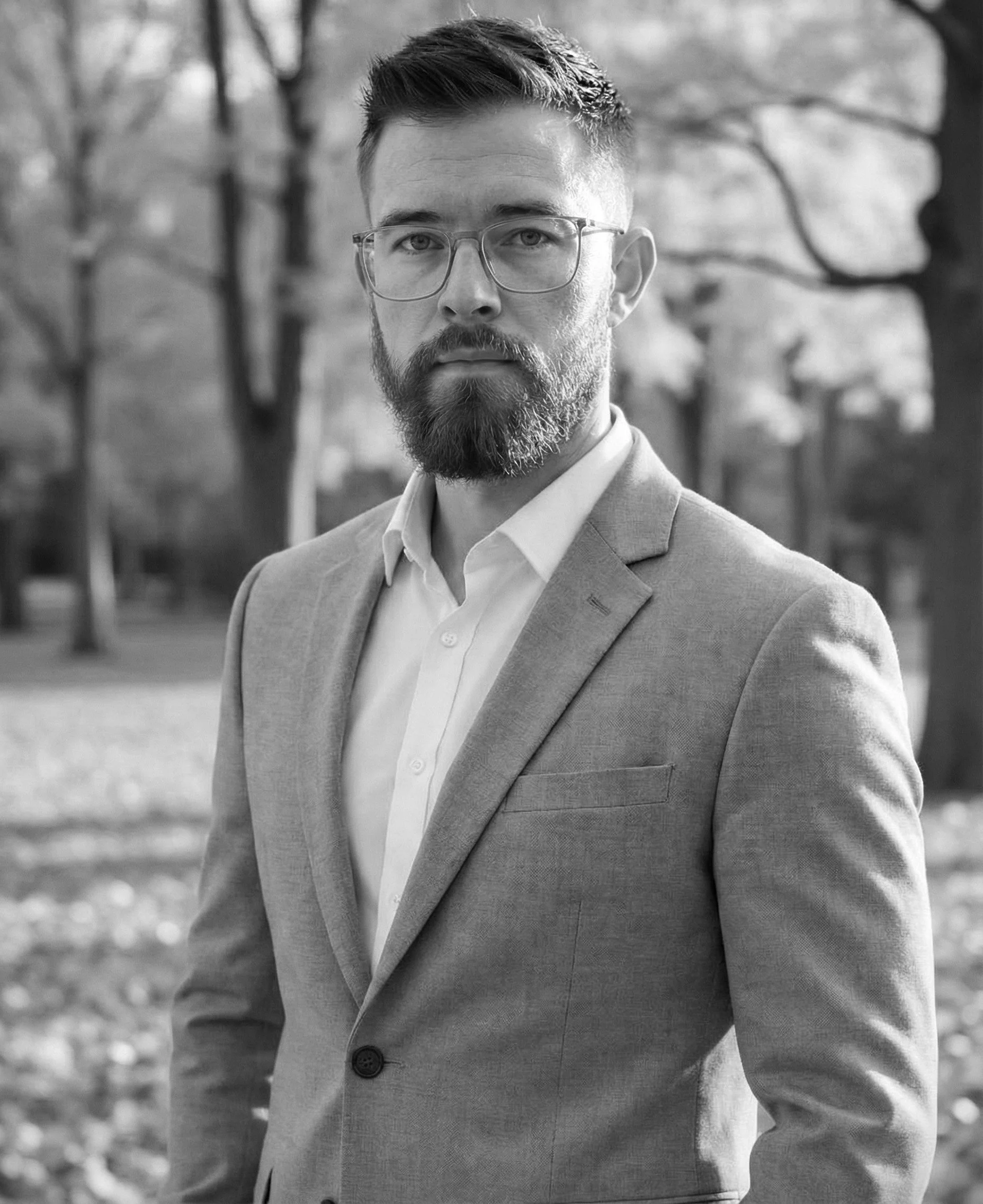 Black and white portrait of a man with glasses, a beard, and short hair wearing a suit jacket and white shirt, standing outdoors with trees in the background.