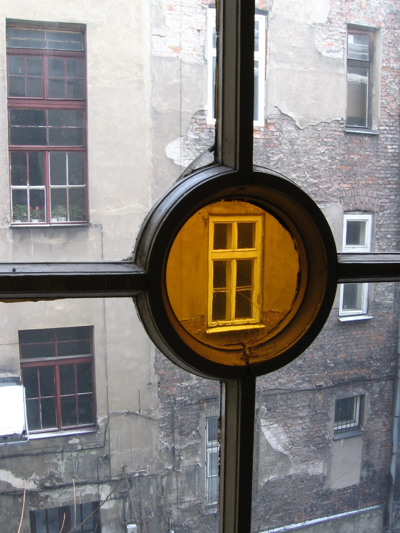 Janela de vidro com moldura de metal, refletindo uma janela antiga de parede de tijolos ao fundo, onde se vê uma janela de quatro painéis. A parede ao redor da janela é de tijolos expostos e parede descascando. Cracóvia, Polônia.