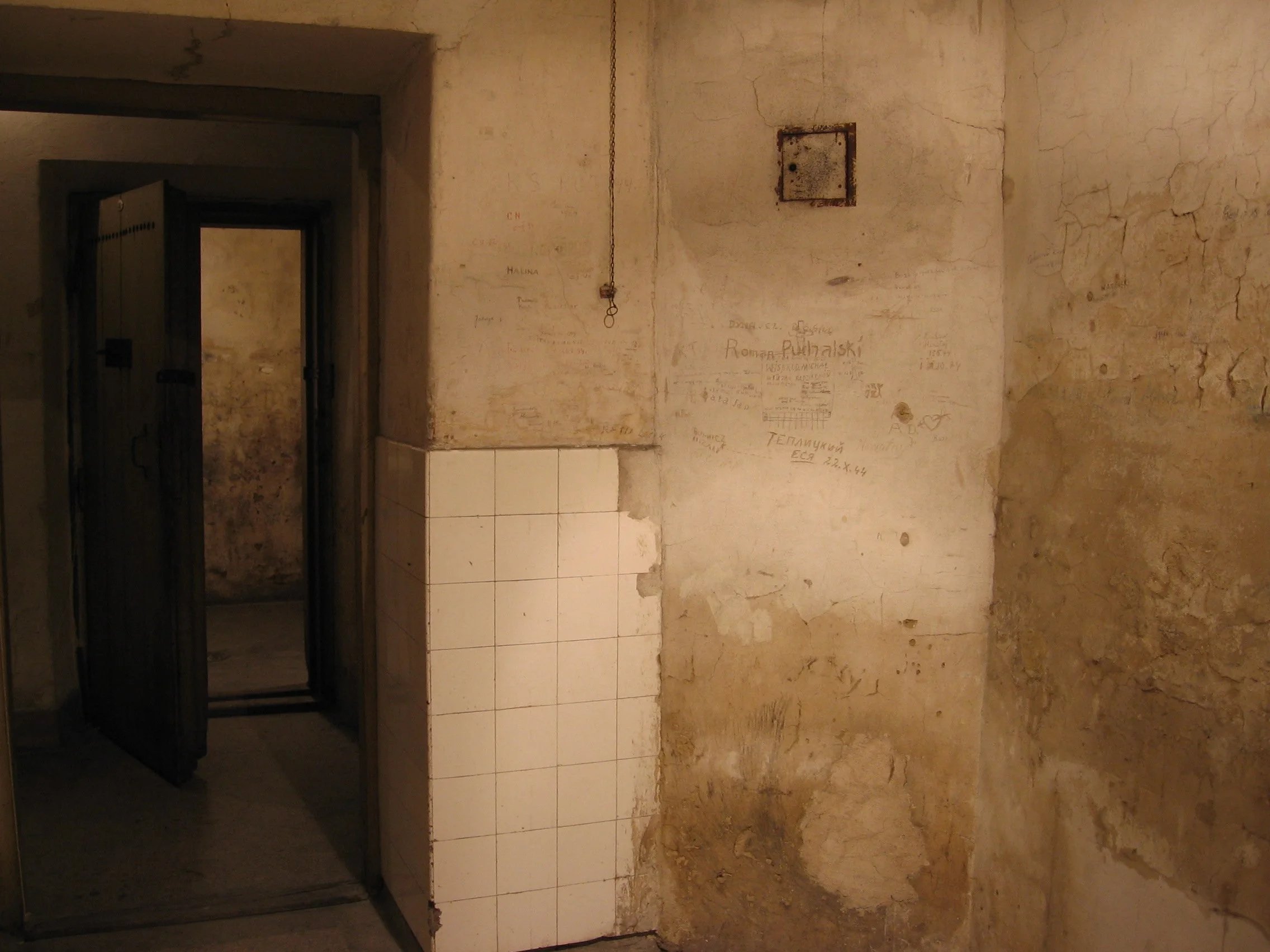Parede rústica e envelhecida com tinta descascada, marcações e graffiti, ao lado de uma porta de ferro aberta, levando a um corredor escuro. Cela de um prédio da Gestapo em Cracóvia, Polônia. 