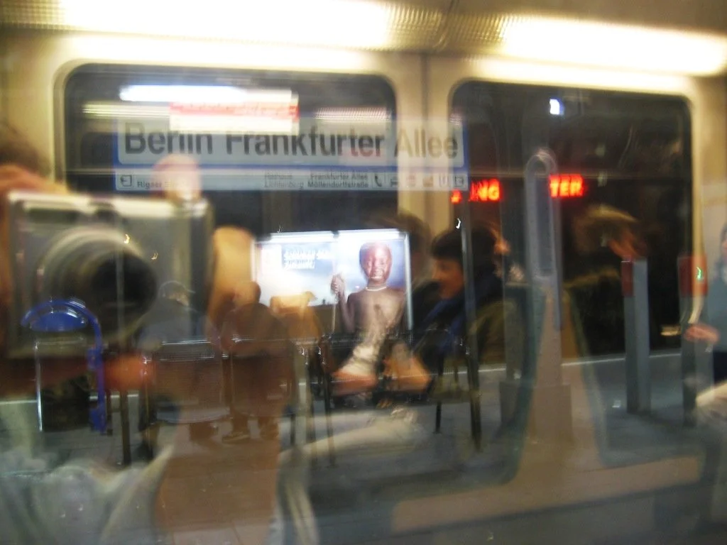 Visão através da janela de uma estação de metrô mostrando um painel com o nome 'Berlin Frankfurter Allee', pessoas sentadas e reflexos de passageiros na janela. Metrô em Berlim. 