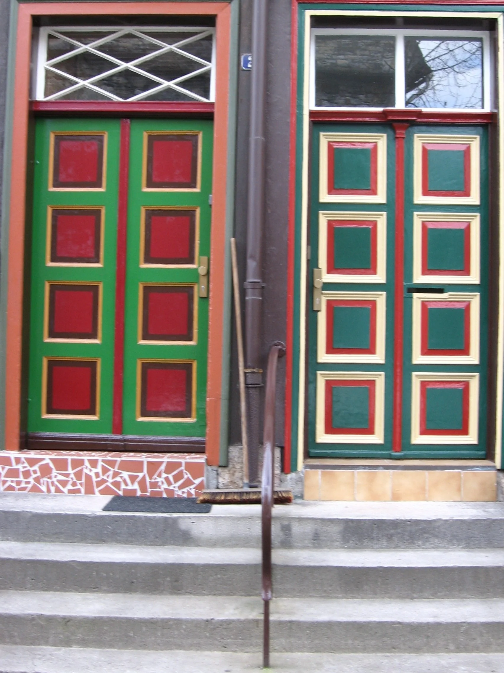 Duas portas de entrada de casas decoradas com cores verdes, vermelhas, amarelas e detalhes em marrom, com degraus de concreto na frente. Casinhas vizinhas em Hannover-Schmündel, Alemanha. 
