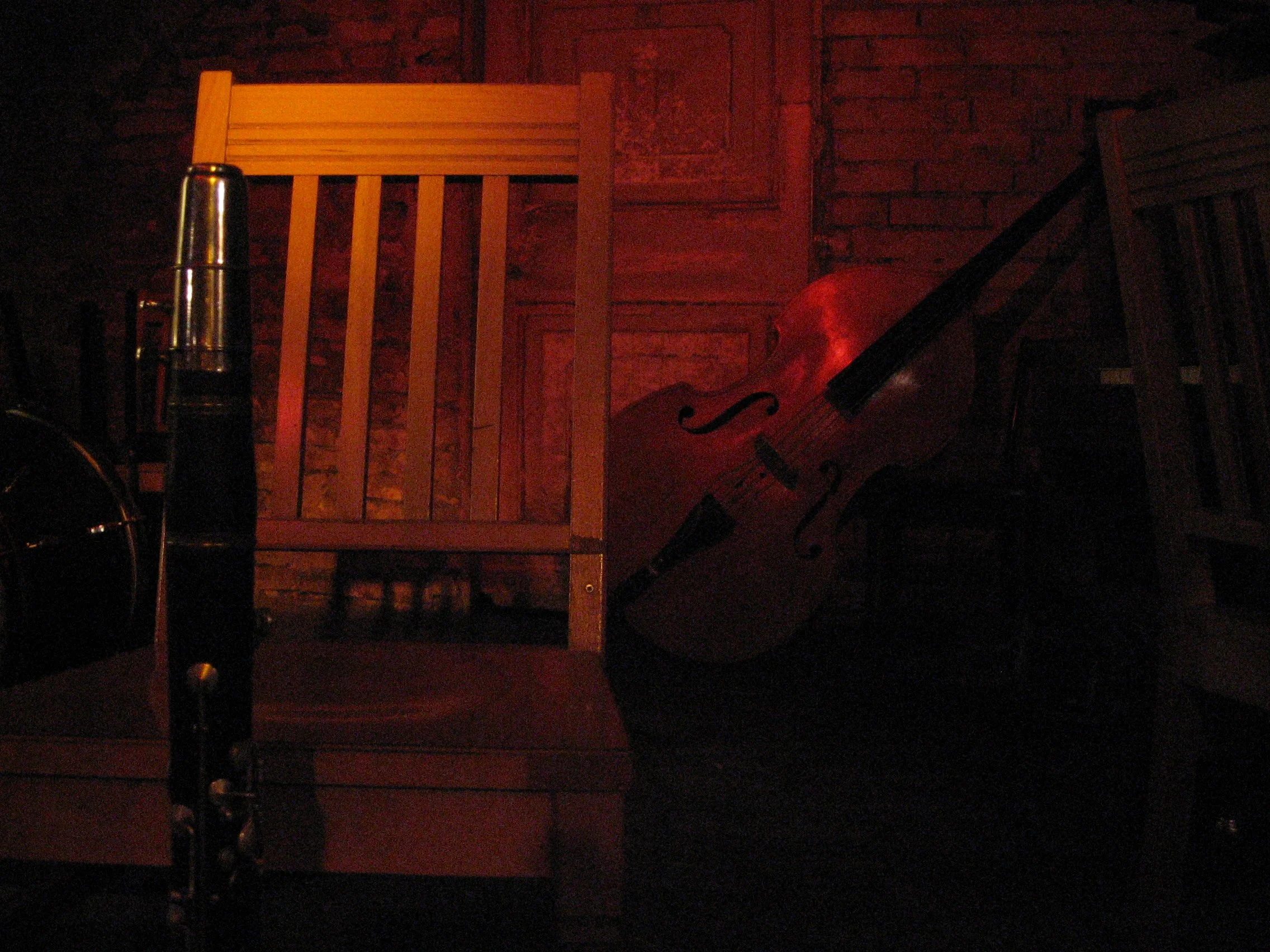 Cena com cadeira de madeira, violino apoiado na cadeira, e um violoncelo encostado na parede de tijolos, com iluminação avermelhada. Bar em Berlim, Alemanha.