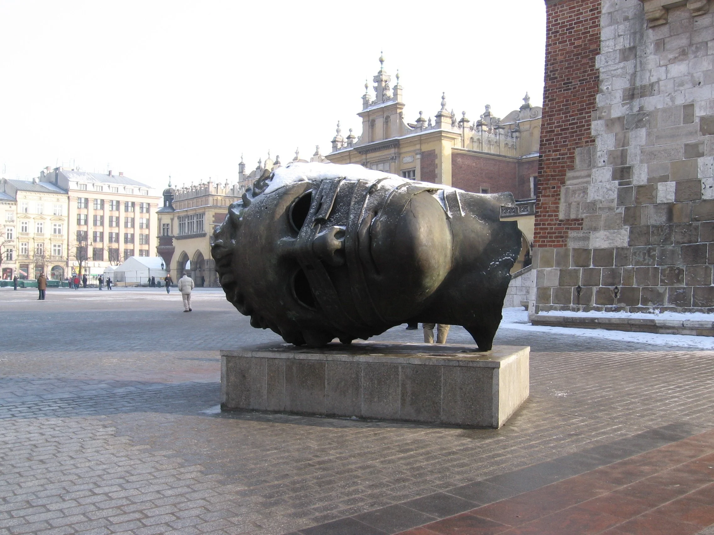 Escultura de cabeça humana de bronze deitada em praça, com edifícios históricos ao fundo. Praça principal de Cracóvia, Polônia. 