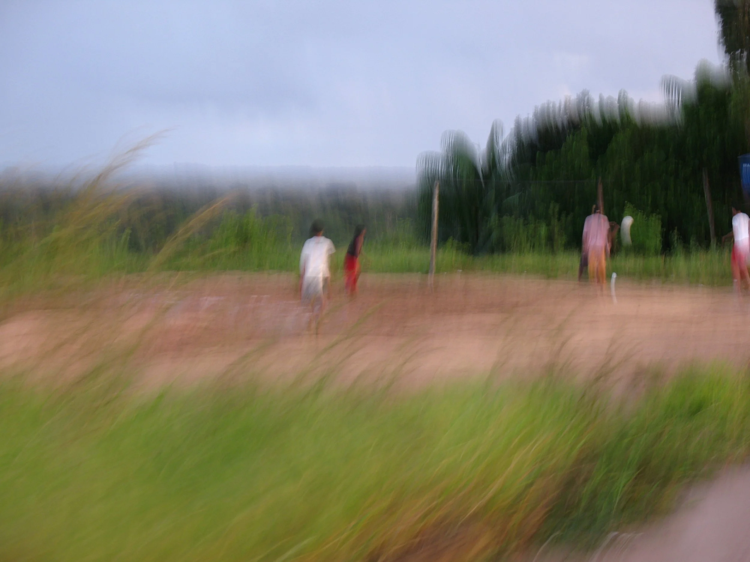 Imagem de uma quadra de vôlei ao ar livre, tendo algumas pessoas jogando, ao fundo árvores e céu nublado, com efeito de movimento ou desfoque. Vila da Moa, perto de Cruzeiro do Sul, Acre.