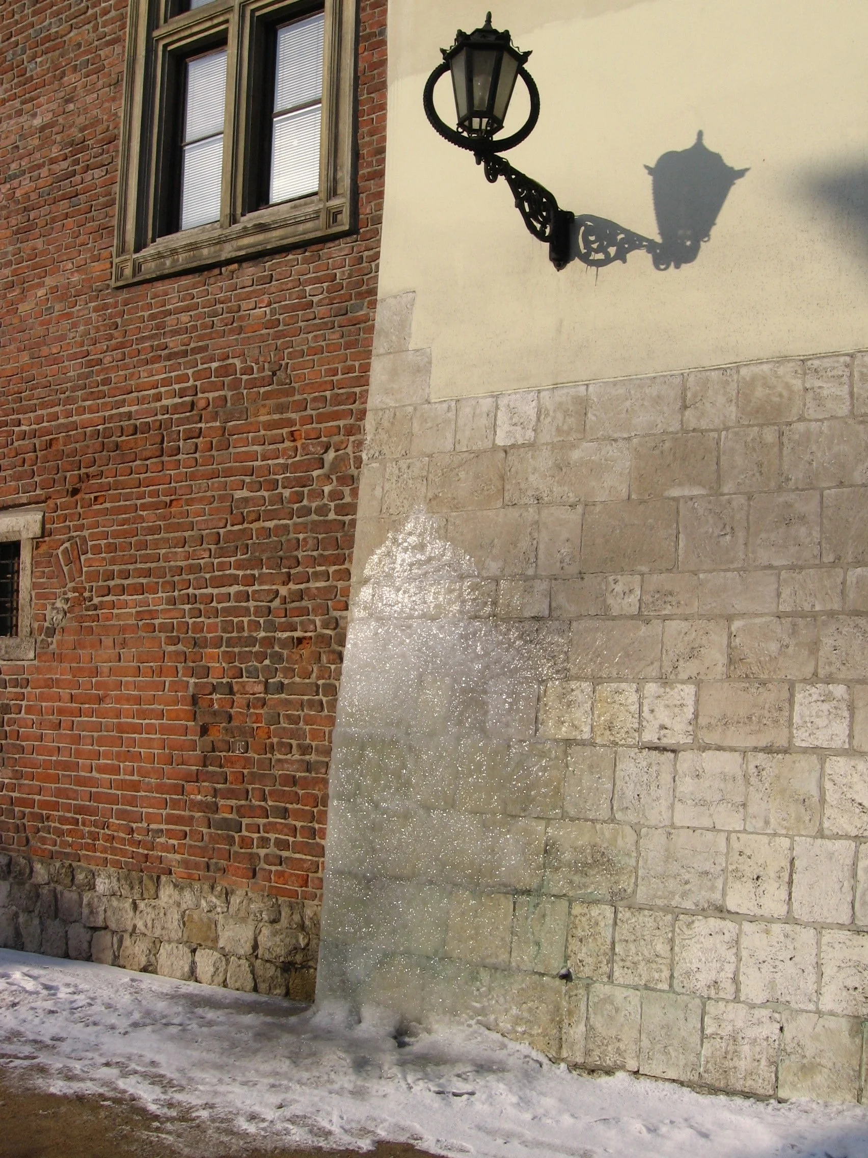 Parede de prédio com uma mistura de tijolos vermelhos e pedras, uma janela com moldura de madeira e uma luminária de rua que projeta uma sombra na parede. Cracóvia, Polônia.