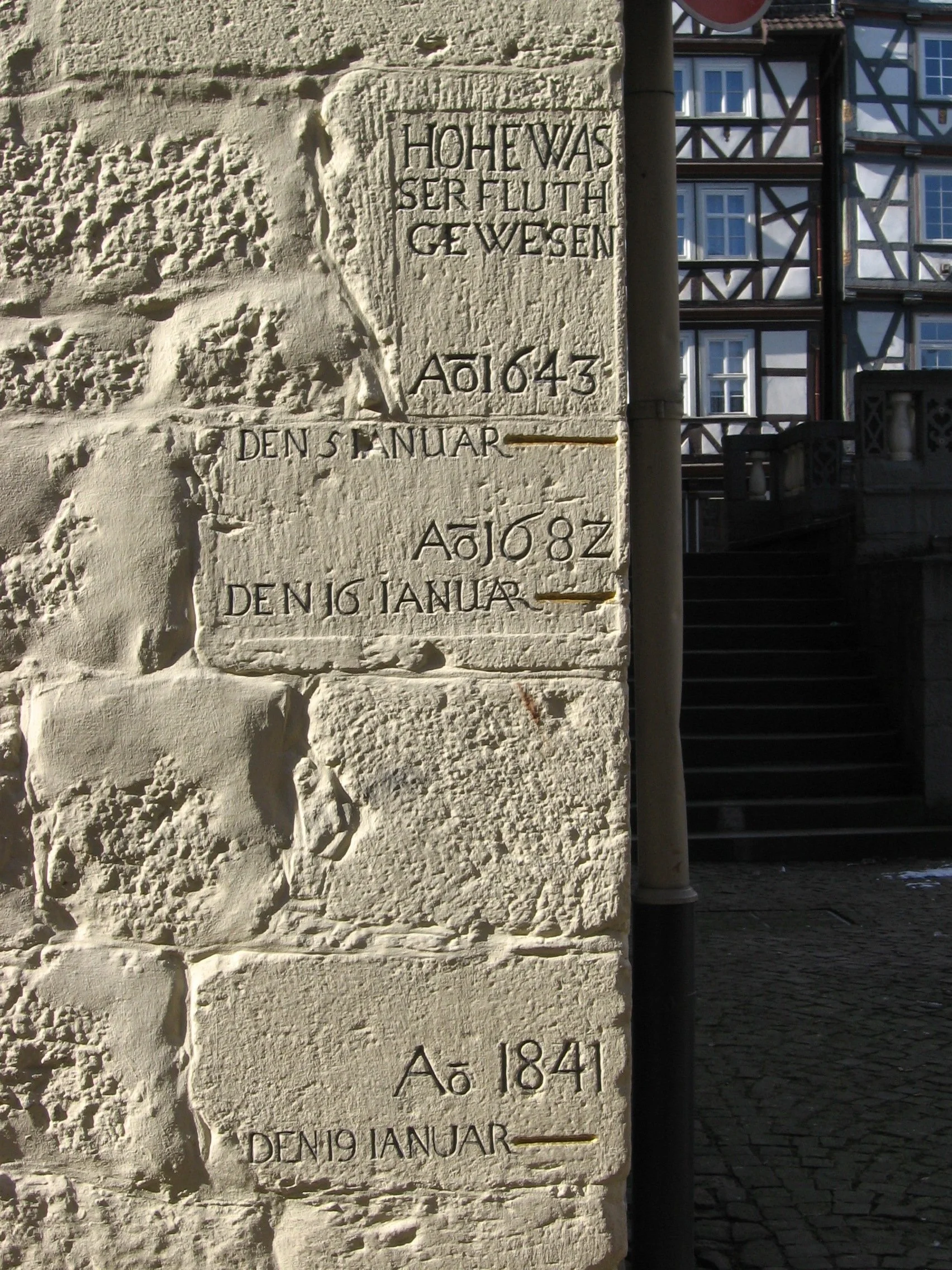 Pedra de monumento com inscrições em alemão, incluindo datas e referências ao rei Luís. Mostra marcas de erosão e parte de um cenário urbano ao fundo, com escadaria e construções de madeira. Marcas de nível de enchente em Hannover-Schmündel, Alemanha