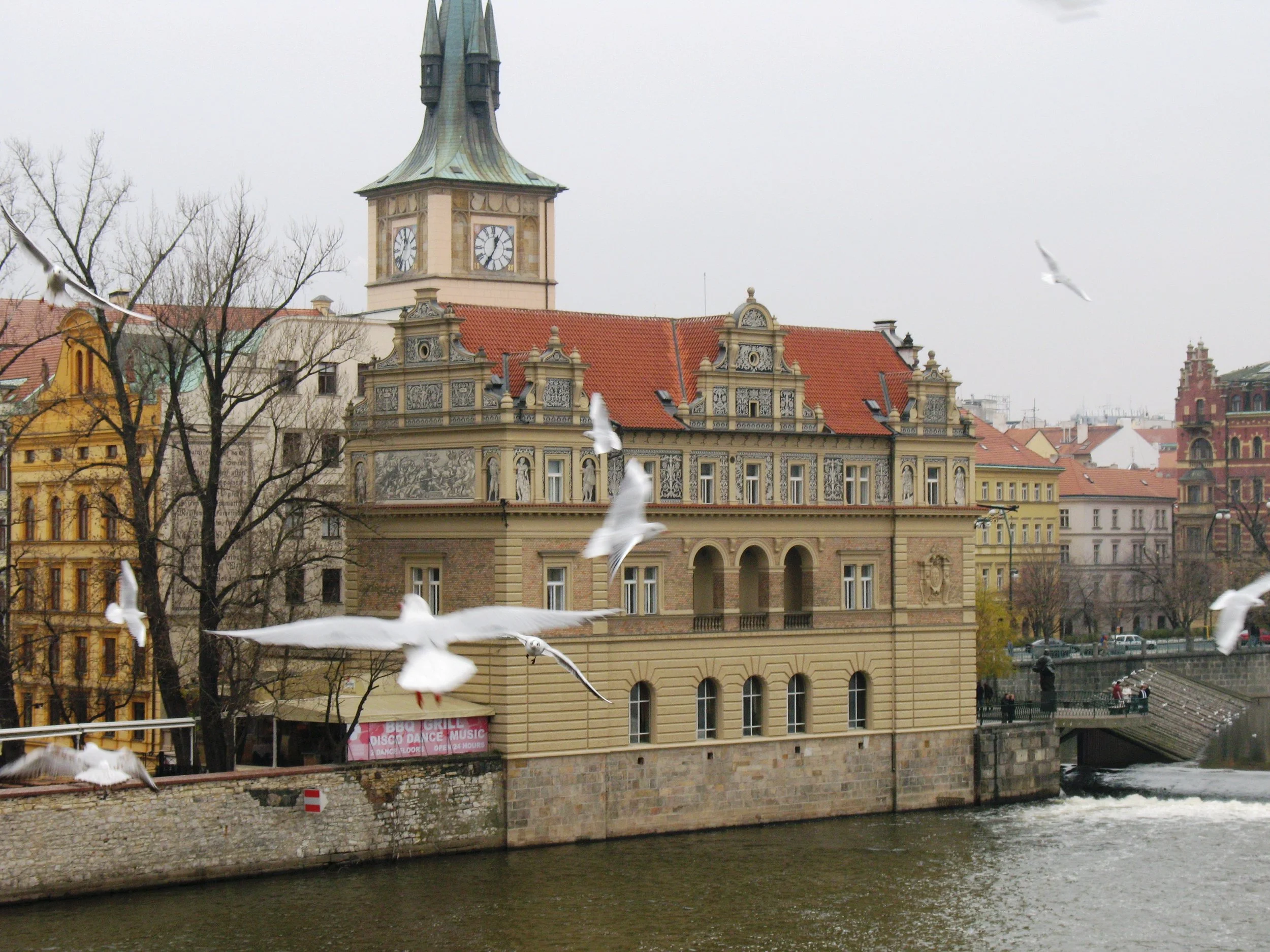 Vista de edifícios históricos ao lado de um rio, com pássaros voando, árvores sem folhas e céu nublado em uma cidade europeia. Praga, República Tcheca. 