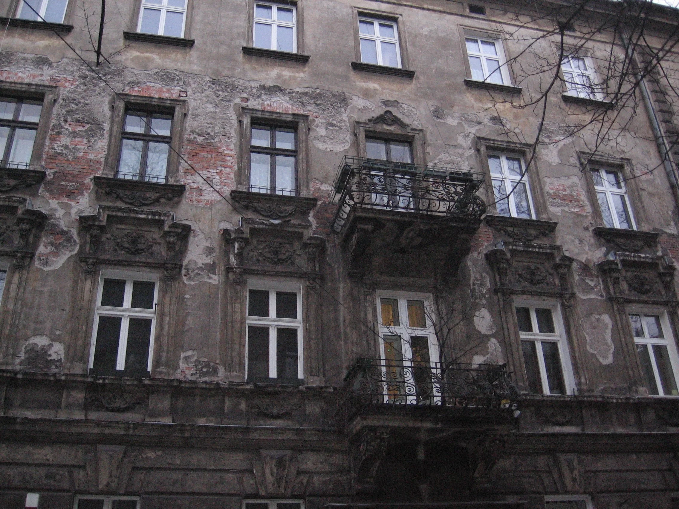 Prédio antigo com janelas de vidro e uma varanda de ferro. A fachada do prédio mostra sinais de desgaste e reboco descascando, com áreas de tijolos expostos. Em Cracóvia, Polônia.