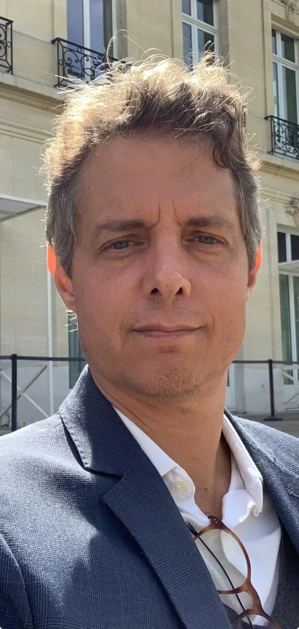 Retrato de um homem de cabelo castanho claro, barba por fazer, vestindo um terno cinza e camisa branca, em frente a um prédio da OCDE em Paris, com janelas e sacadas de ferro. Alexandre Constantino.