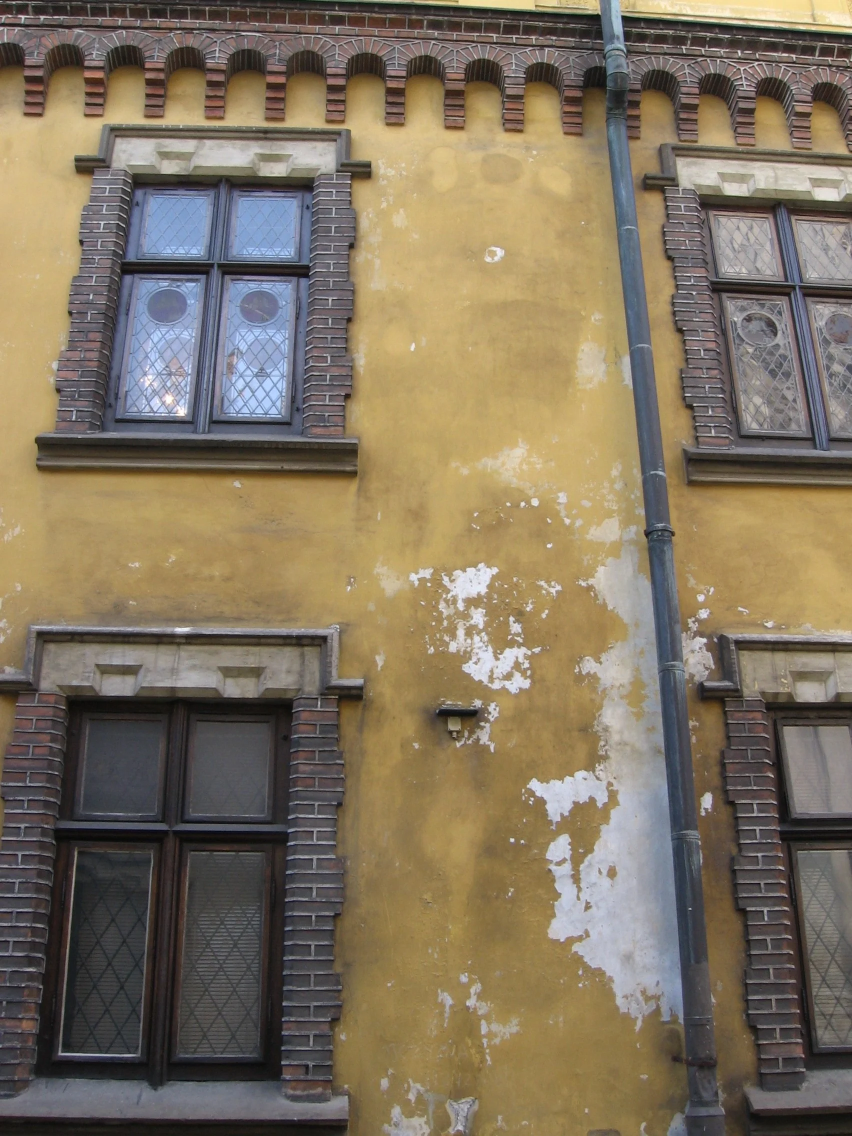 Fachada de prédio antigo com paredes amarelas desgastadas, janelas com caixilhos de madeira e molduras de pedra, detalhes em tijolos na volta das janelas e uma calha de chuva na lateral. Cracóvia, Polônia.