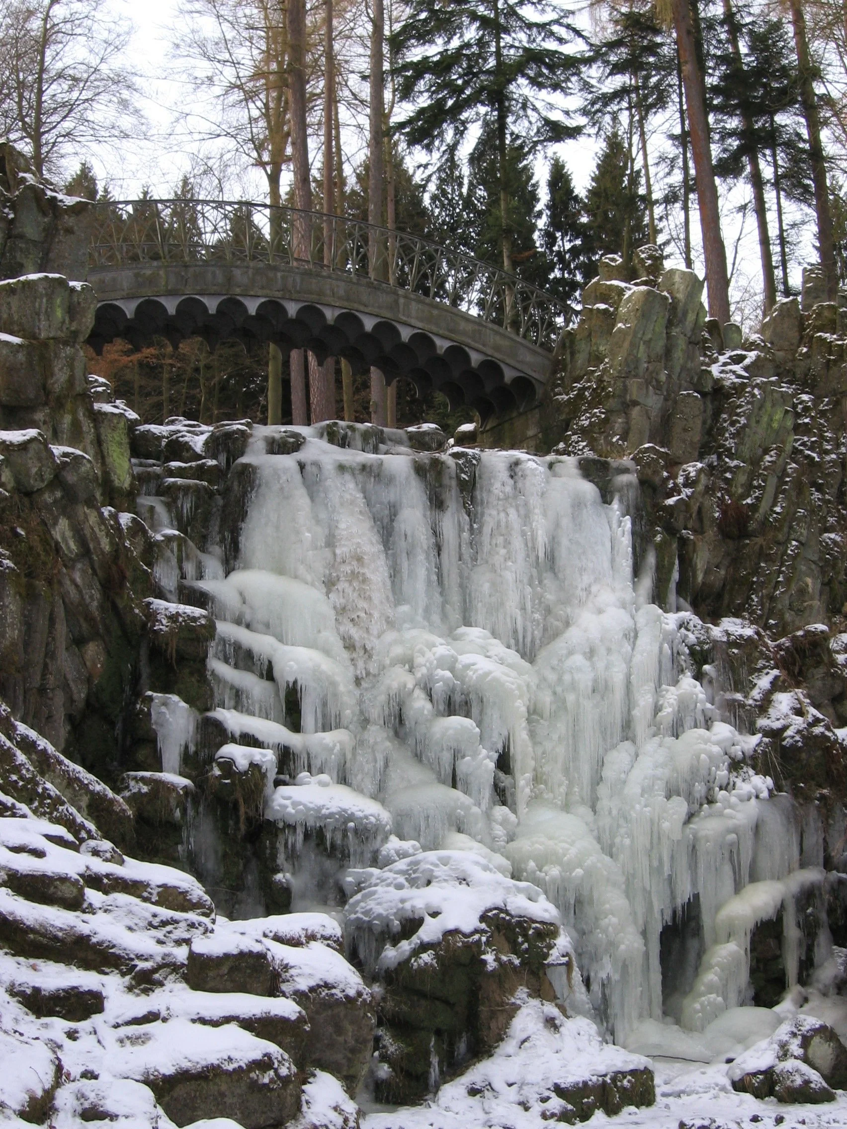 Cachoeira congelada com gelo em rochas ao redor, árvores altas ao fundo e uma ponte de ferro acima. Schloss Wilhelmshöhe, Kassel, Alemanha.