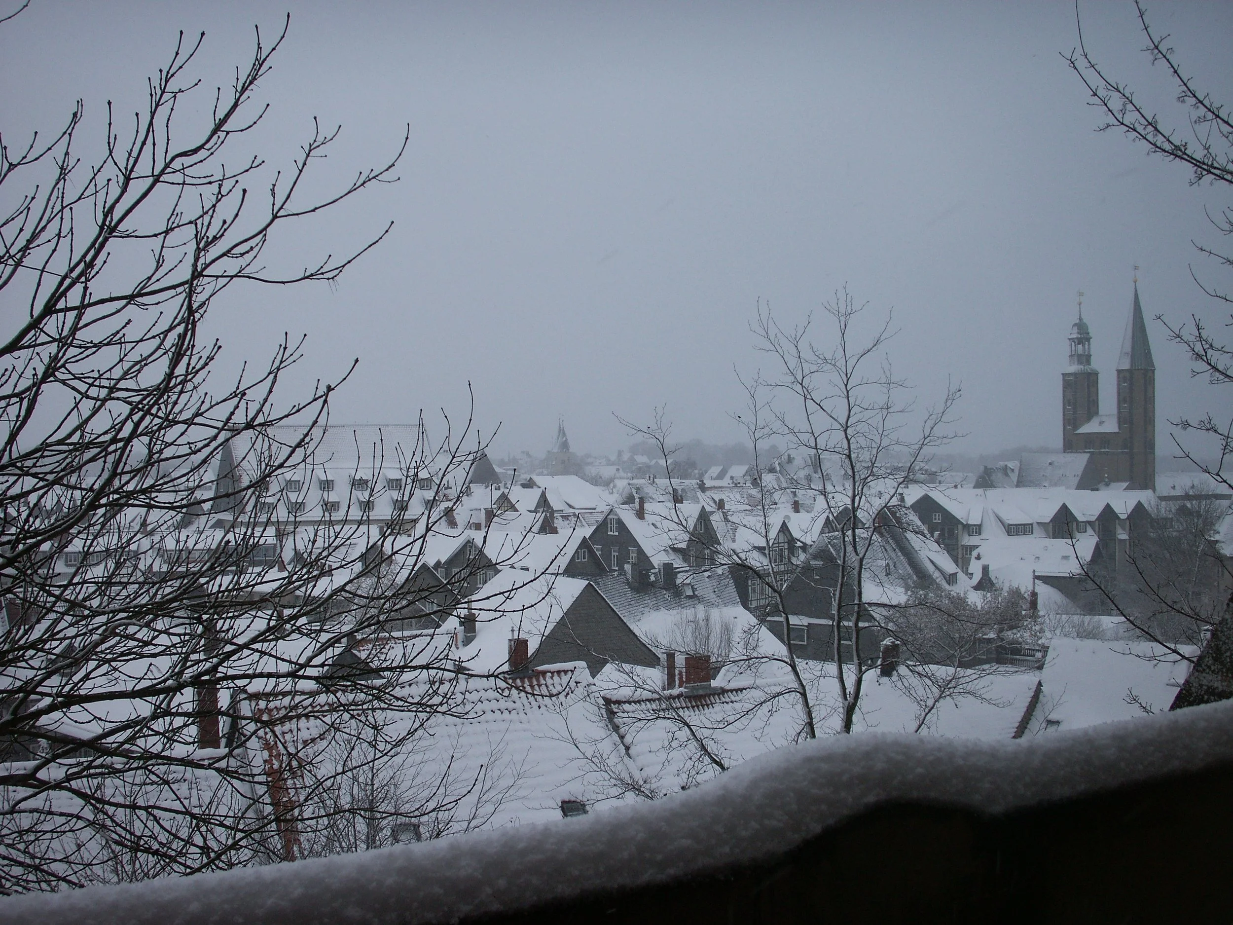 Cena de uma cidade coberta de neve, com telhados nevosos, árvores sem folhas na frente e uma igreja grande ao fundo, tudo sob um céu nublado. Kaiserpfalz Goslar, Alemanha.
