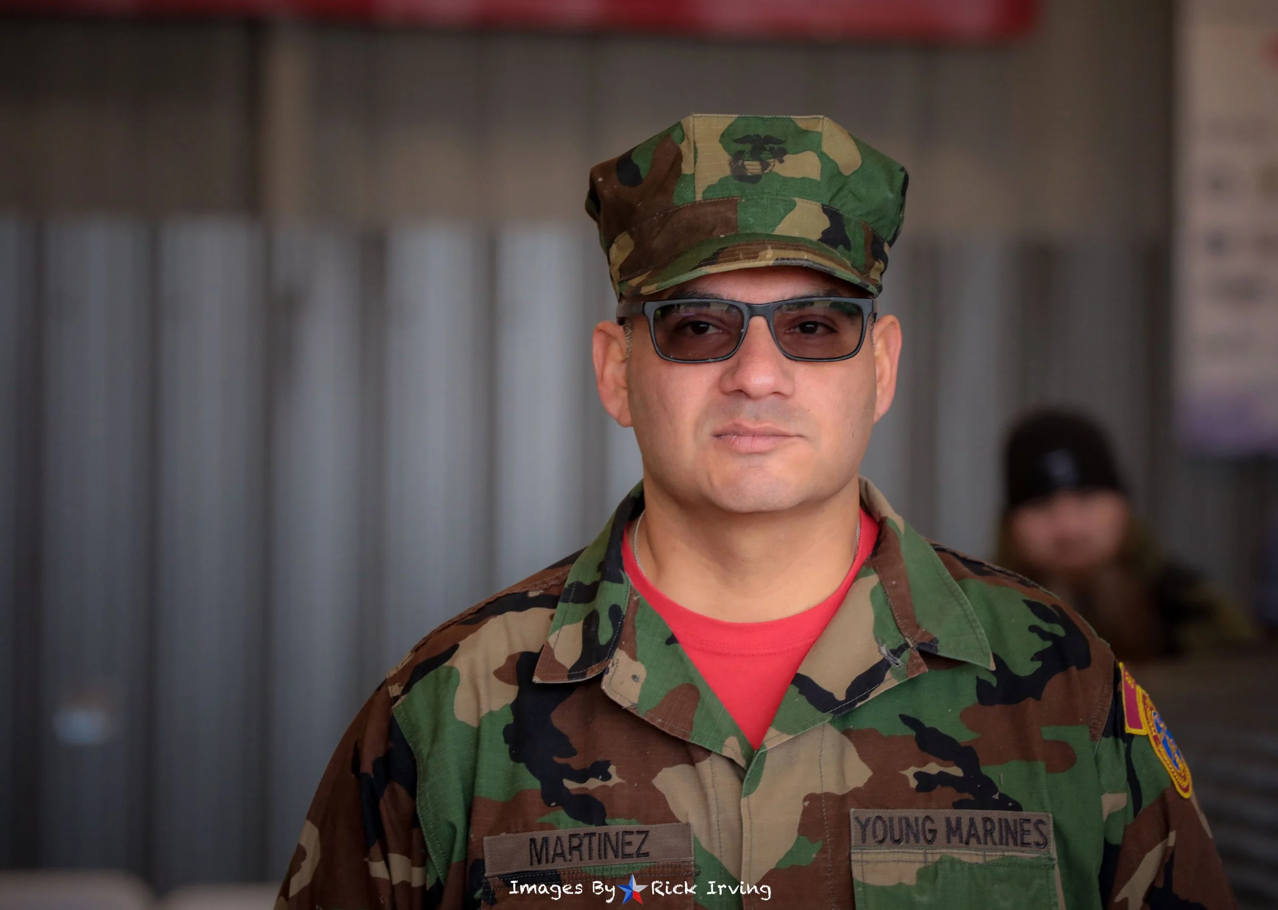 A man in military uniform with camouflage pattern, wearing glasses, standing indoors. His uniform has a name tag that says 'Martinez' and an additional patch that reads 'Young Marines'.