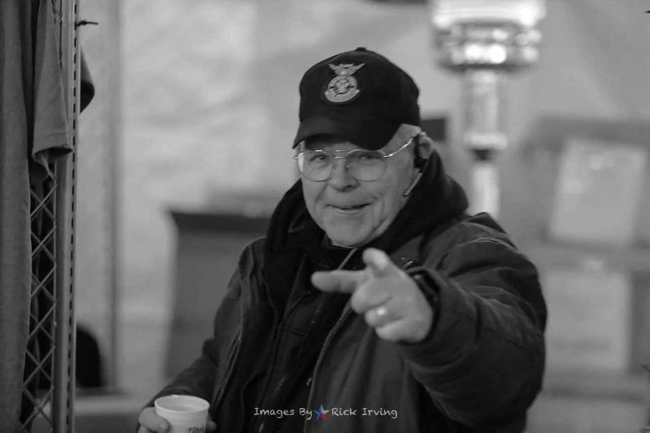 A man wearing glasses, a baseball cap with a Marine Corps emblem, and a jacket, pointing towards the camera while holding a cup, in a setting that appears to be indoors.