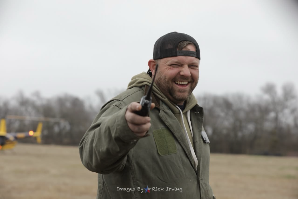 Smiling man in a baseball cap and jacket holding a walkie-talkie outdoors, with a helicopter in the background during overcast weather.