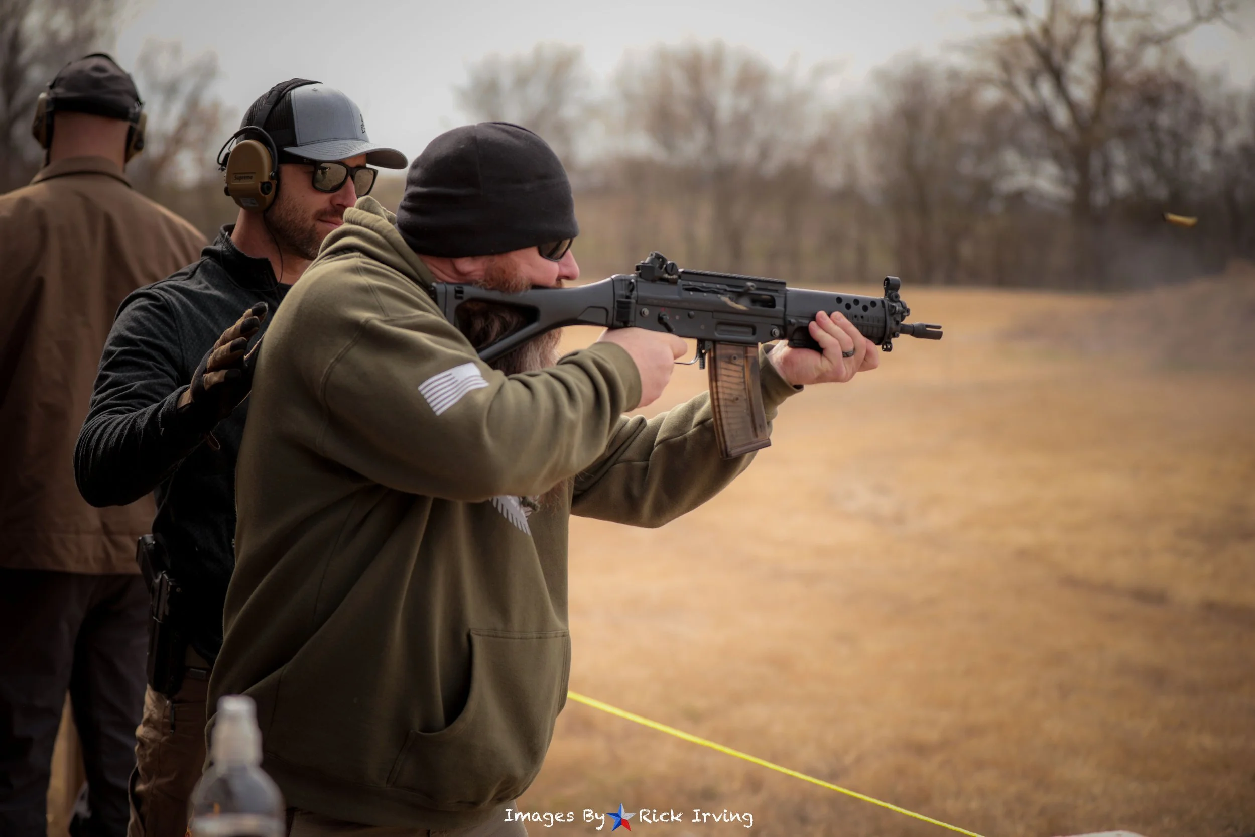 Man in olive green hoodie shooting a rifle at outdoor shooting range with other people nearby.