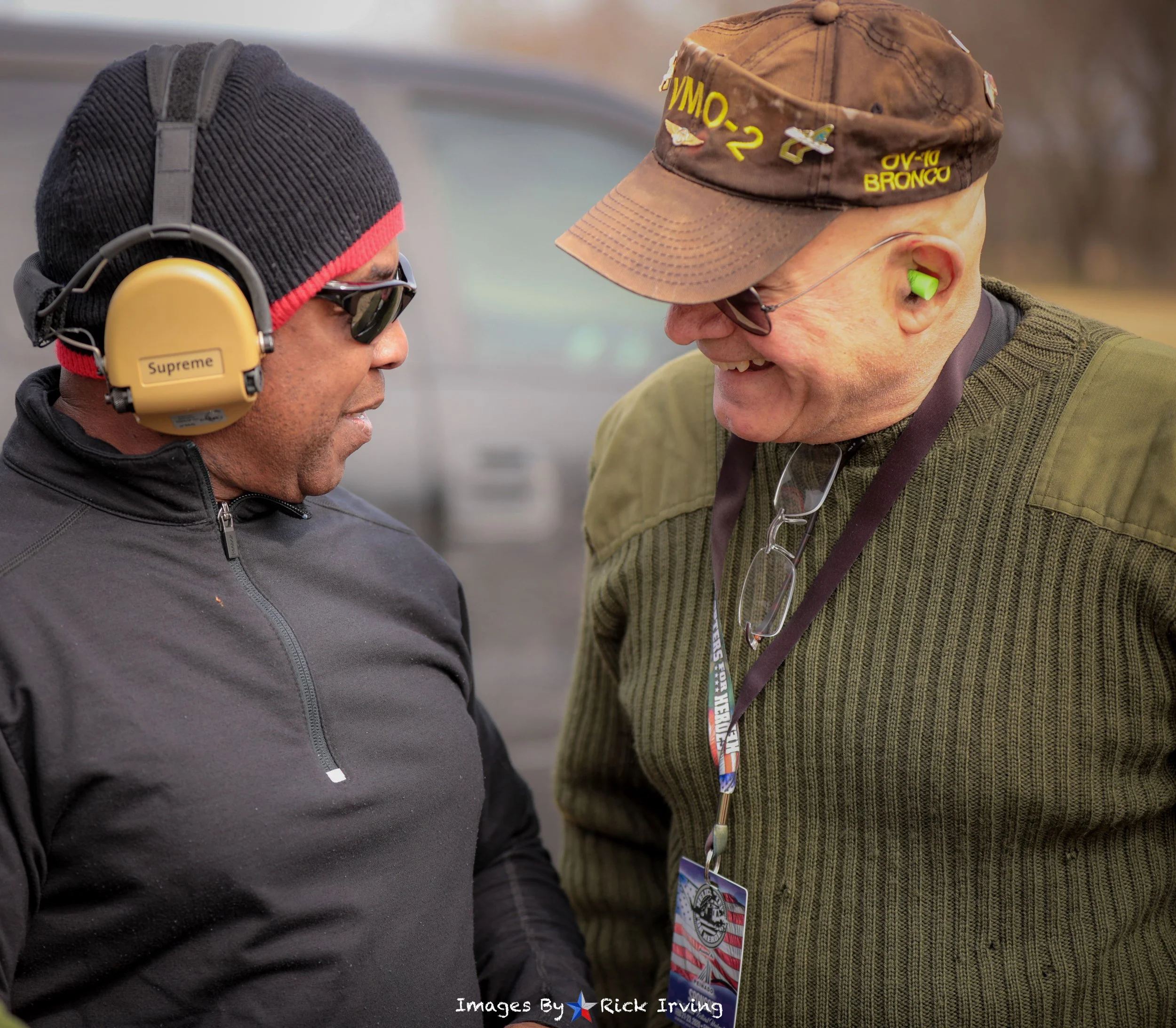 Two men outdoors smiling and talking, one wearing a black jacket, headphones, and sunglasses, the other in a green sweater, glasses hanging around neck, camouflage hat with pins, and green earplugs.