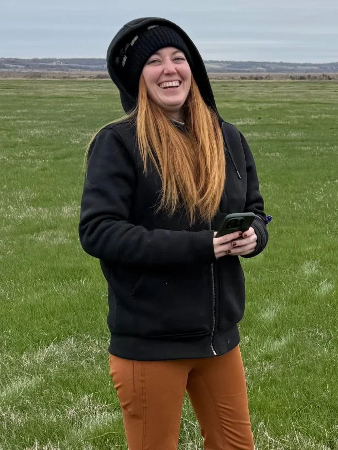 Smiling woman with long red hair, wearing a black beanie, black hoodie, and brown pants standing outdoors on a grassy field and holding a smartphone.