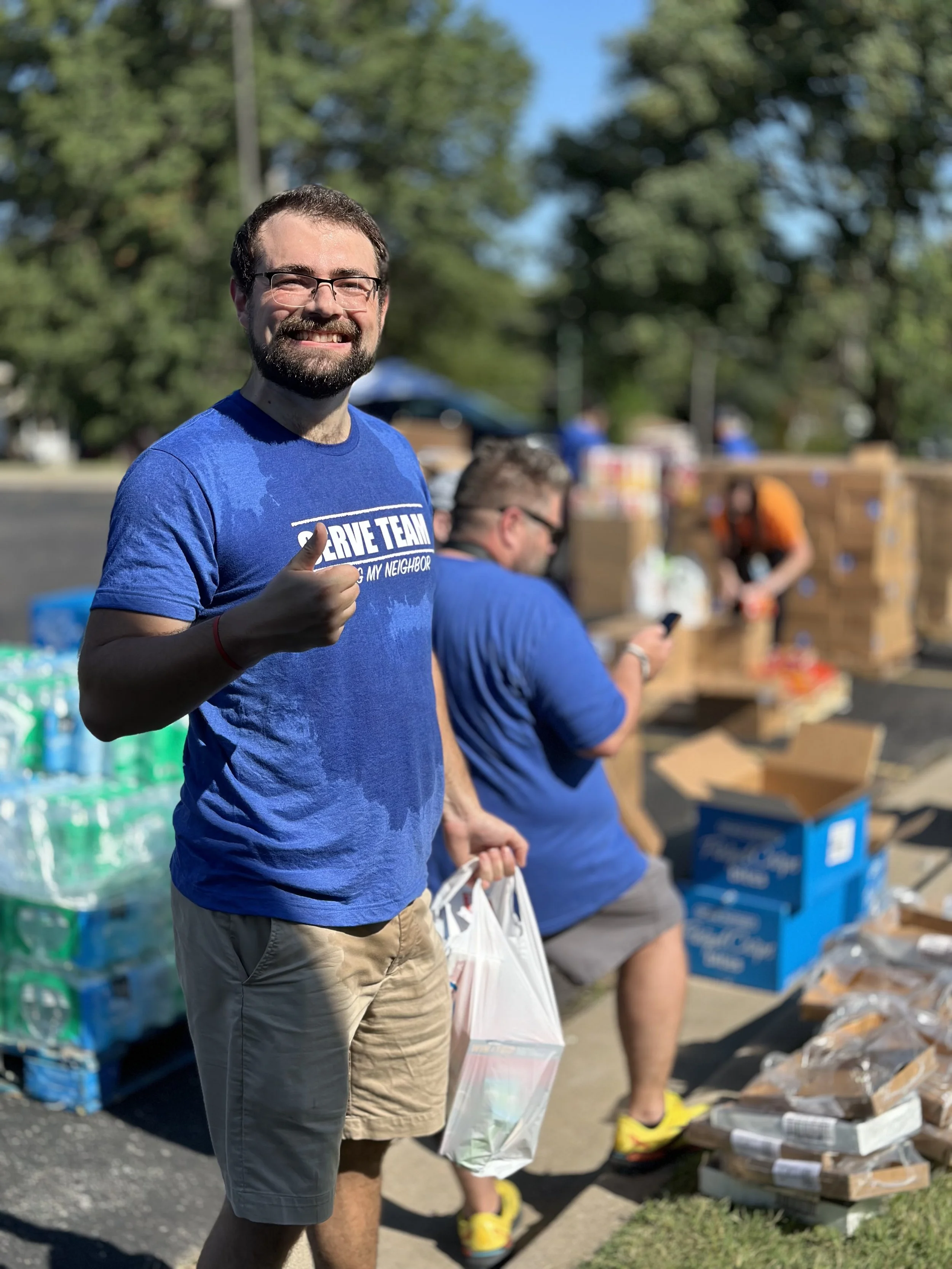 A man in a blue shirt giving a thumbs-up at a community volunteer event with people sorting and packing supplies in the background.