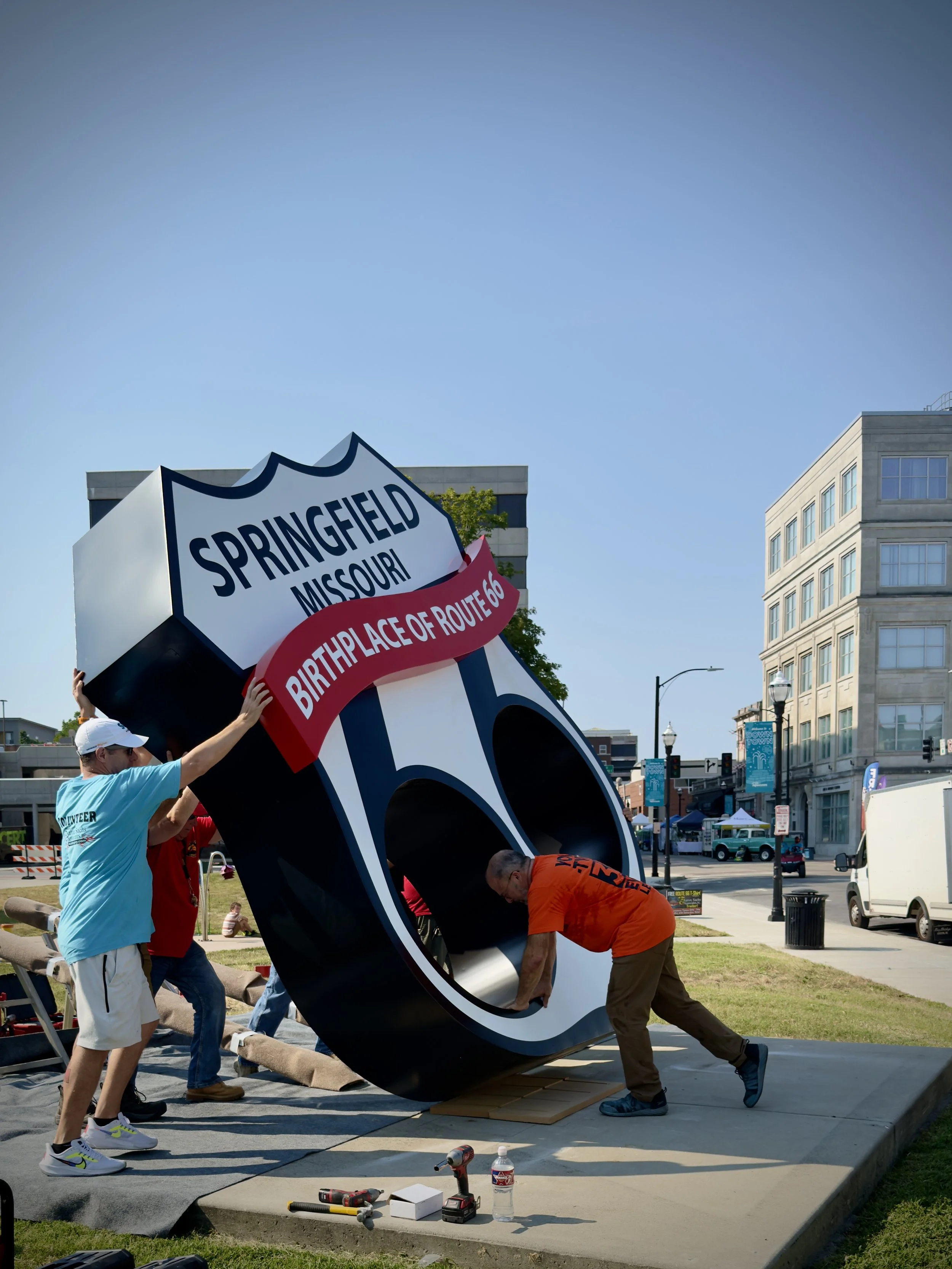 People assembling a large sign in the shape of a highway shield that reads 'Springfield Missouri' and 'Birthplace of Route 66' outdoors in a city park.