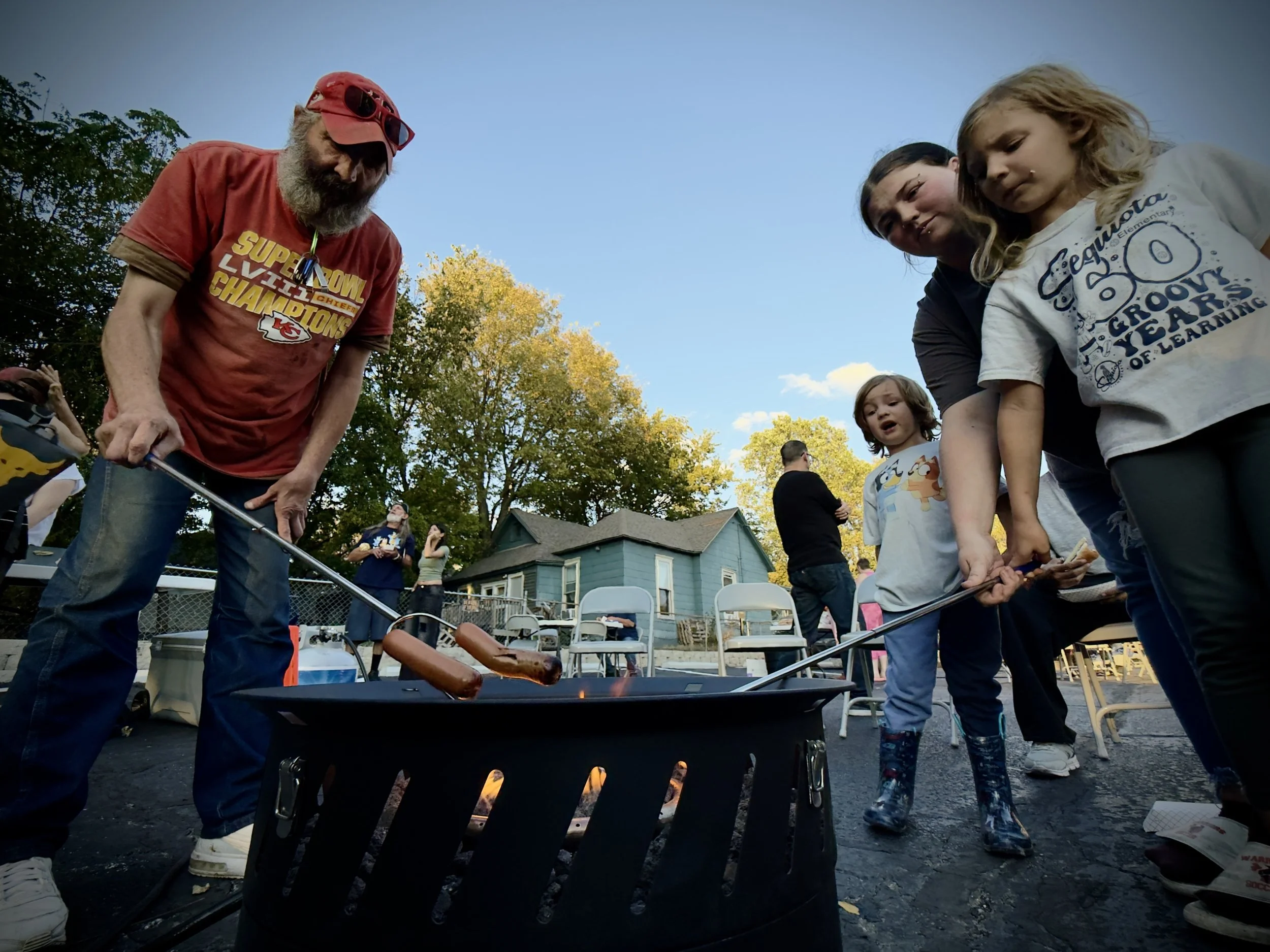 A group of children and adults roasting hot dogs over an outdoor fire pit on a sunny day in a backyard with trees and a house in the background.