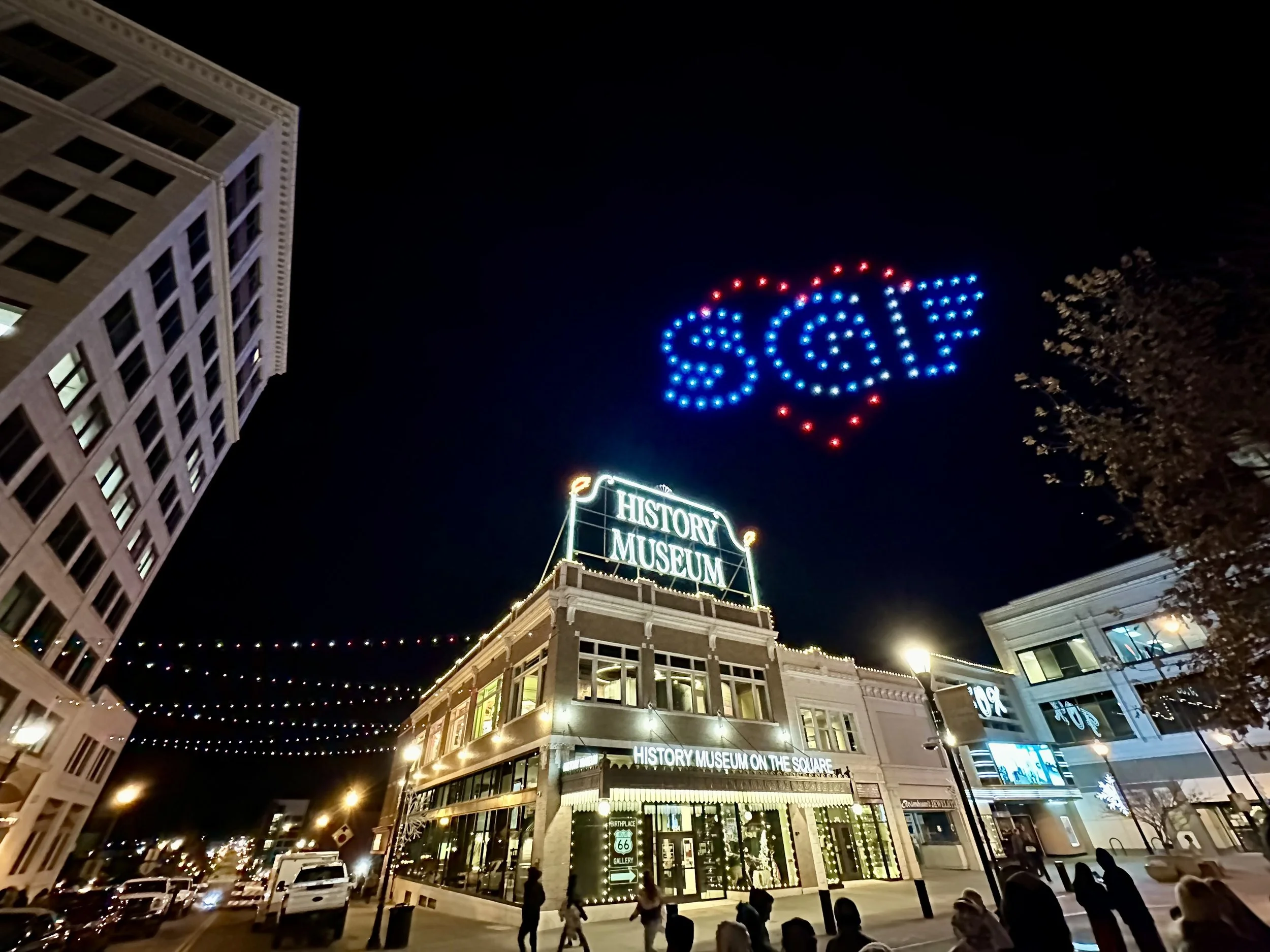 Night view of the History Museum on the Square with festive string lights and a heart-shaped drone light display spelling 'SAY' in the sky.