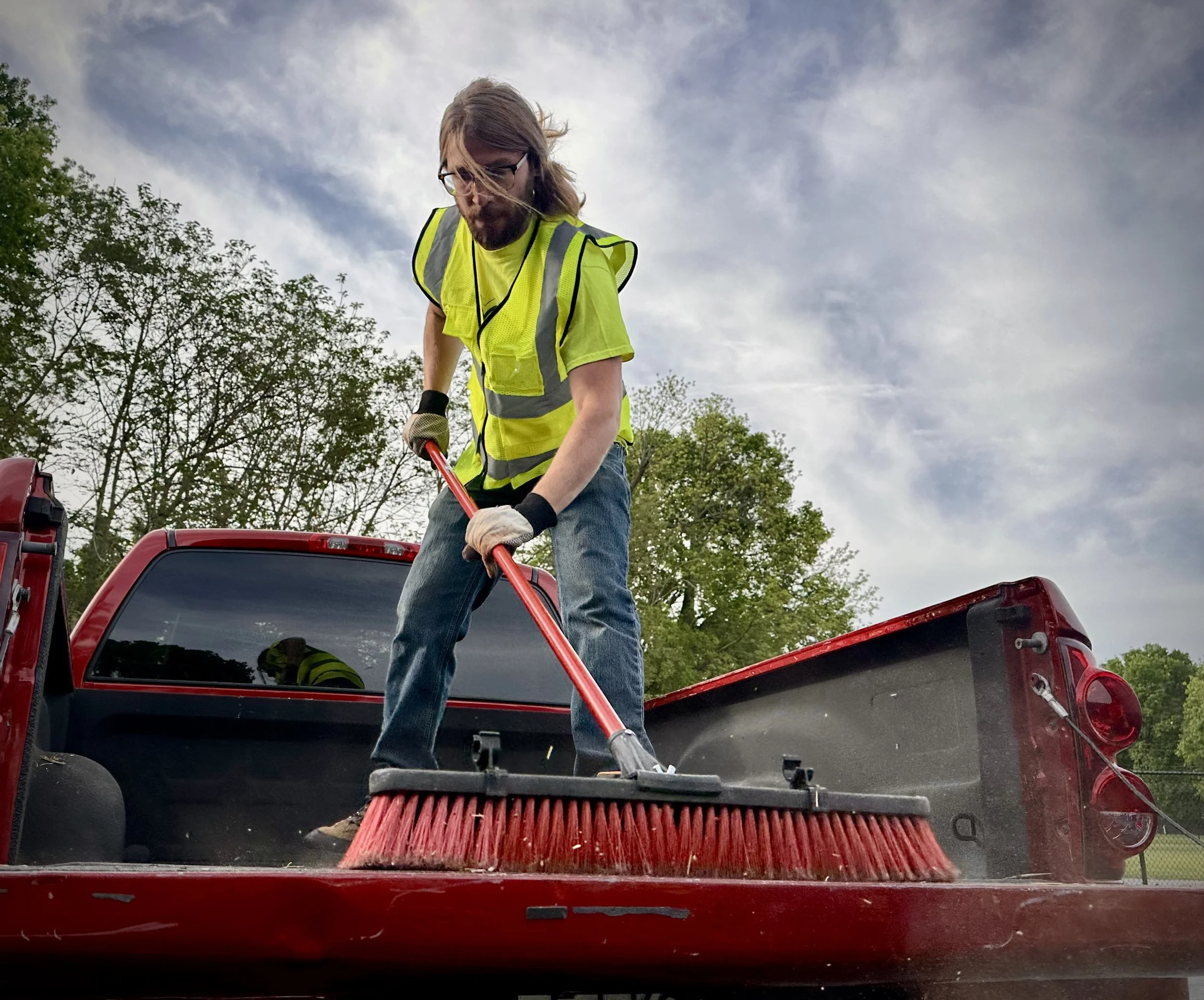 A person wearing a yellow vest and gloves is standing in the back of a red pickup truck using a broom to sweep the truck bed.