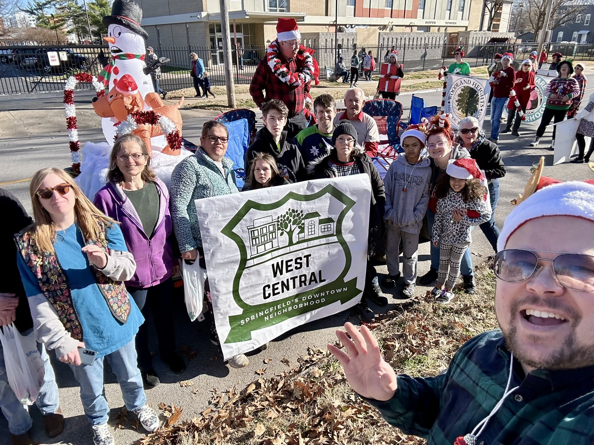 A group of people participating in a holiday parade, holding a banner that reads 'West Central Springfield's Downtown Neighborhood.' They are outdoors, dressed in casual clothing with some wearing Santa hats. There is a decorated snowman figure and festive decorations in the background.