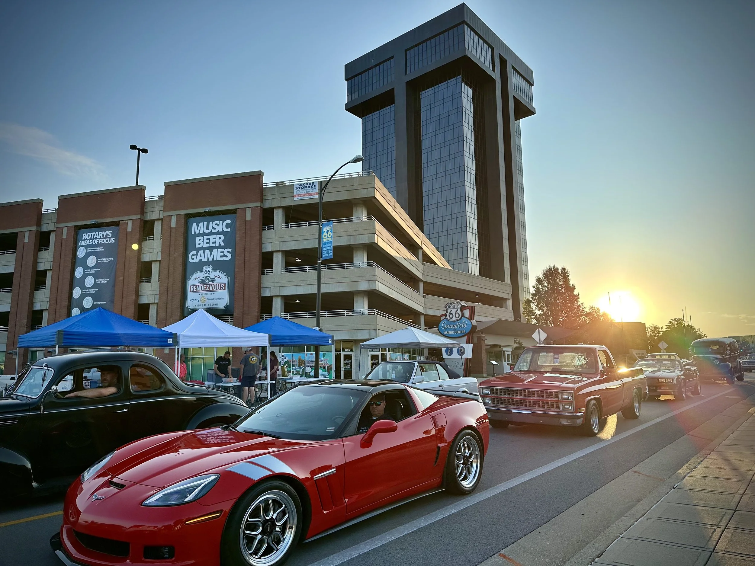 Classic and modern cars parked on street with tents and people outdoors at sunset, in front of a multi-story building with signs for music, beer, and games.
