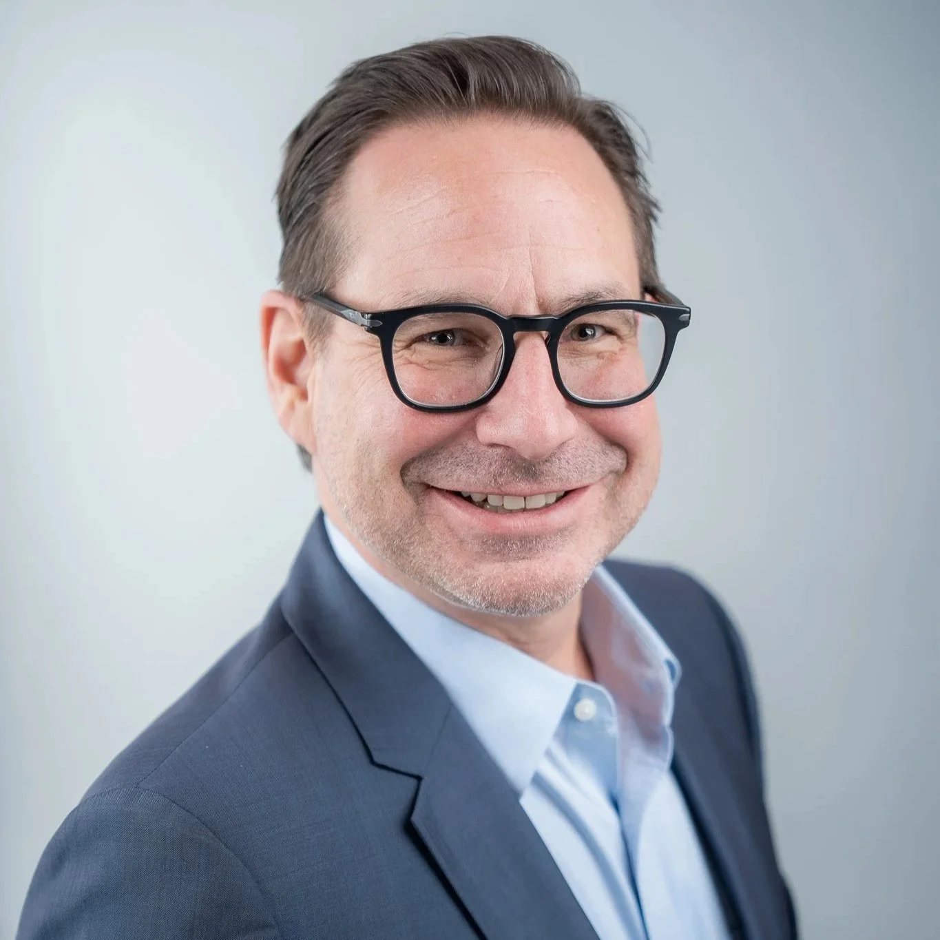A smiling man wearing glasses and a suit in front of a neutral background.