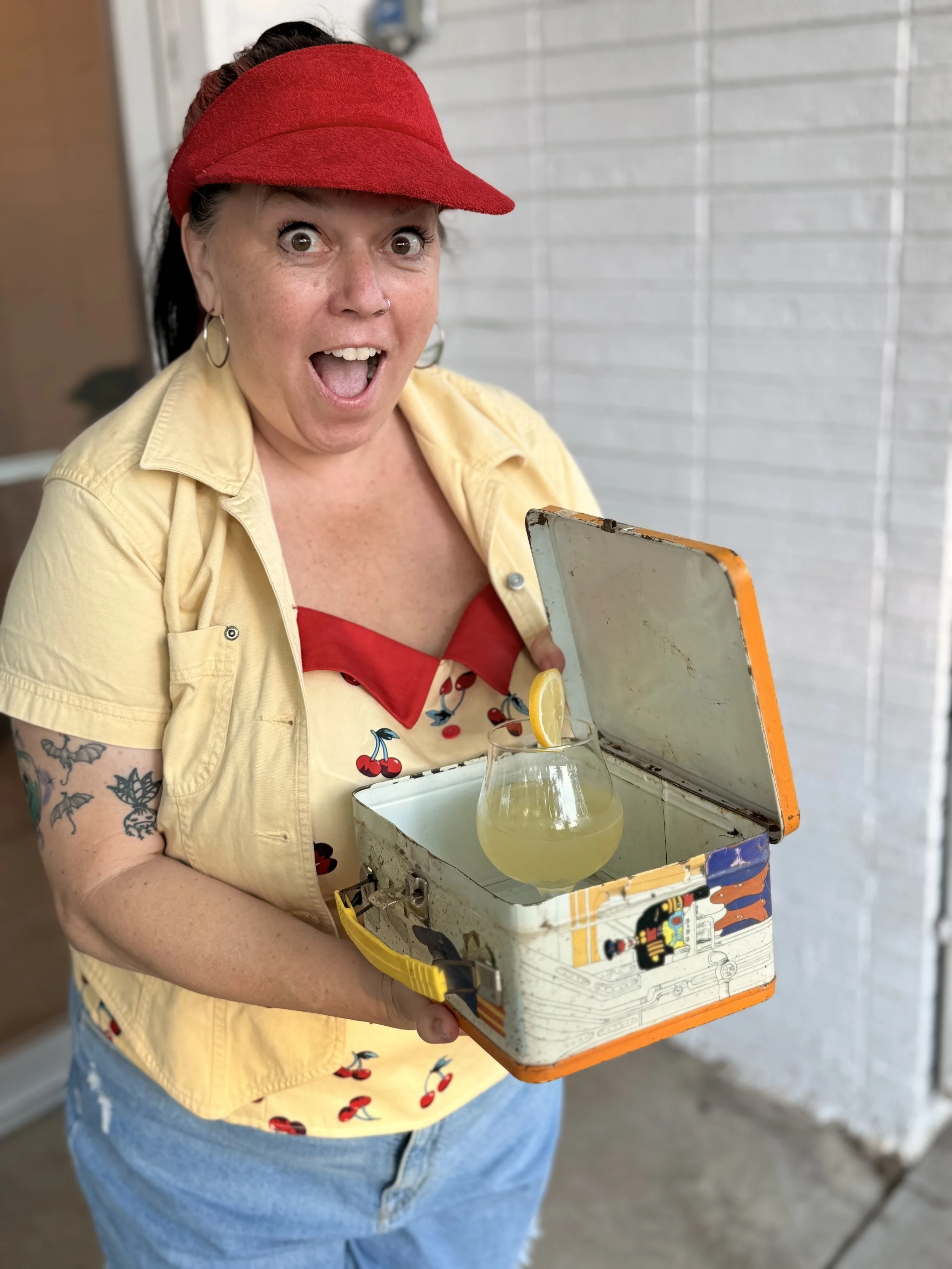 A woman wearing a red visor and a yellow shirt with cherries print, holding a vintage metal lunchbox with a glass of lemonade and a lemon slice, standing in front of a light-colored brick wall, with her mouth open in surprise.