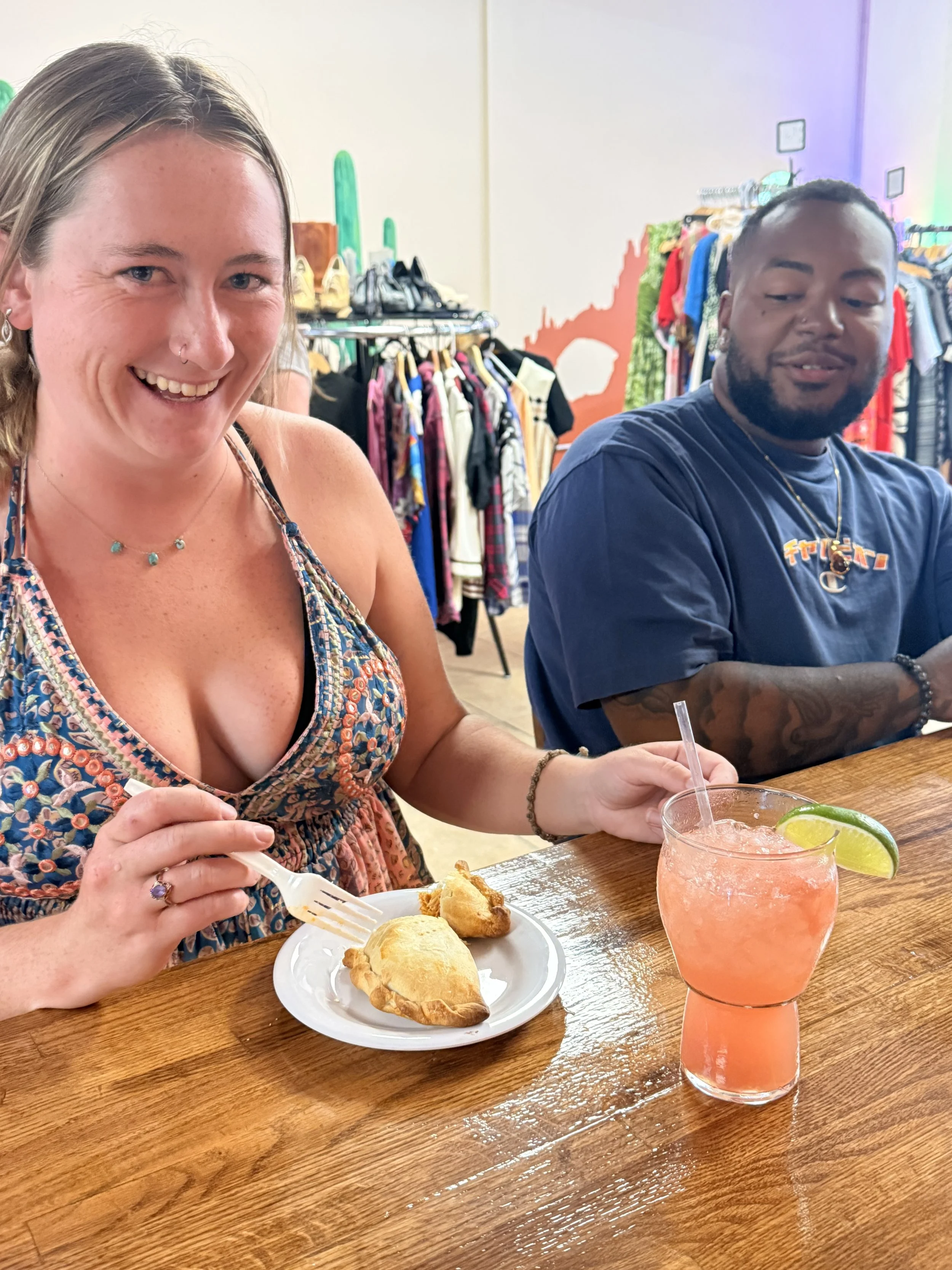 A woman smiling and holding a fork at a restaurant table, with a plate of baked goods and a pink cocktail with a lime wedge in front of her, and a man sitting beside her in the background, inside a clothing store with racks of clothes and shoes.