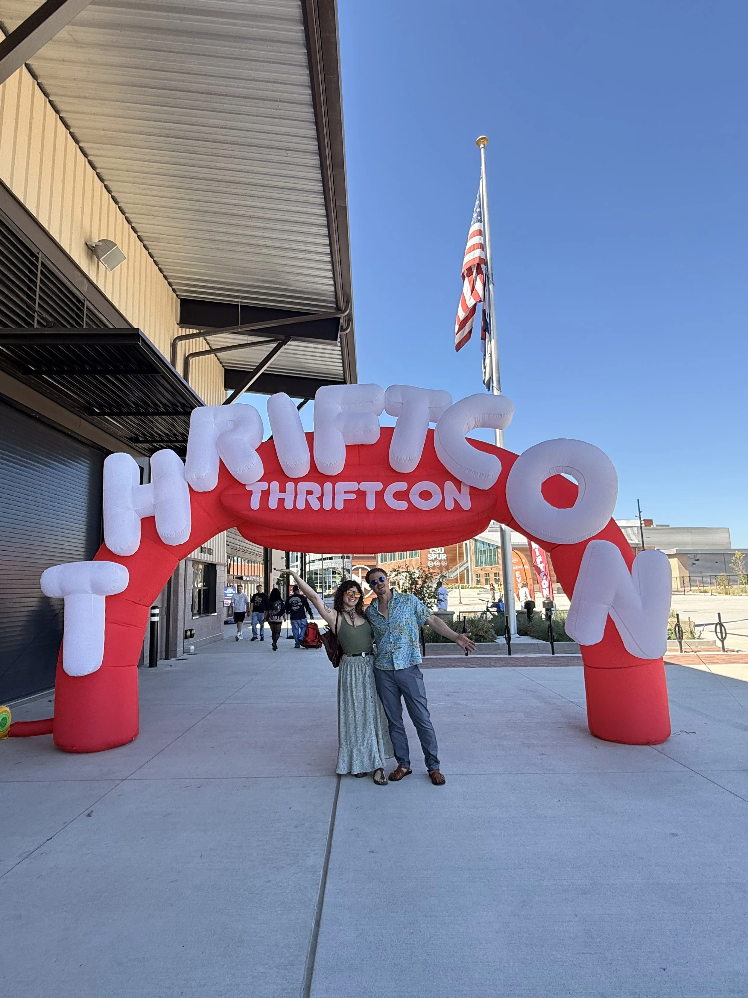 Two people standing under a red inflatable arch with white letters, outside a building on a sunny day. The arch has the words "TRI