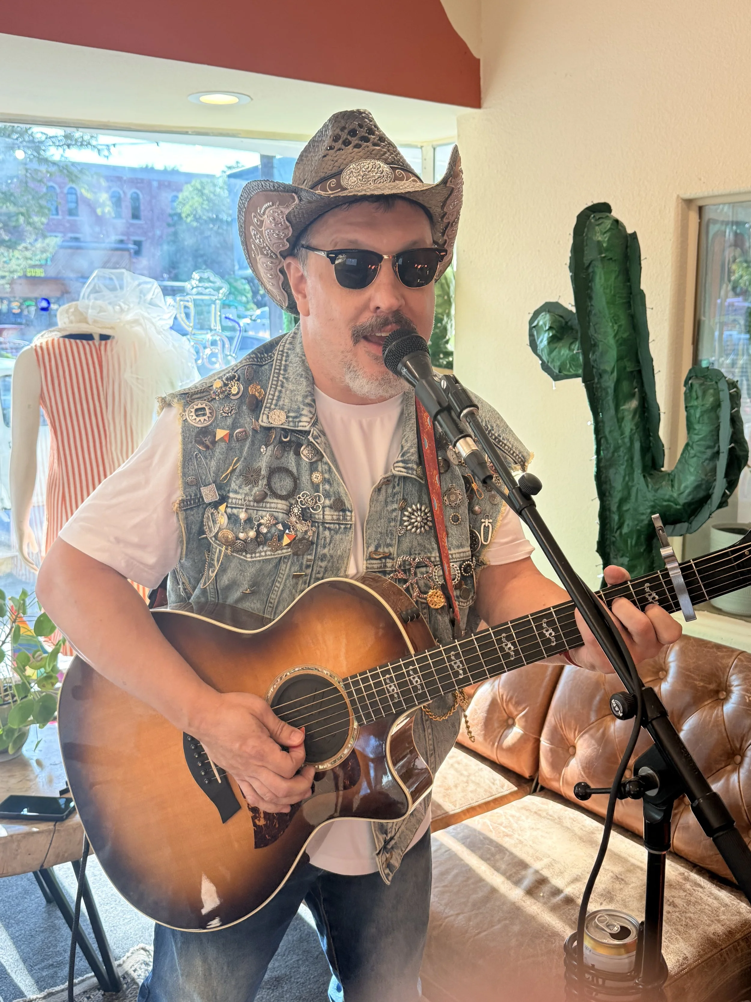 A man wearing a cowboy hat, sunglasses, and a decorated vest is playing an acoustic guitar and singing into a microphone. He is inside a room with a window, a sofa, and various decorative items.