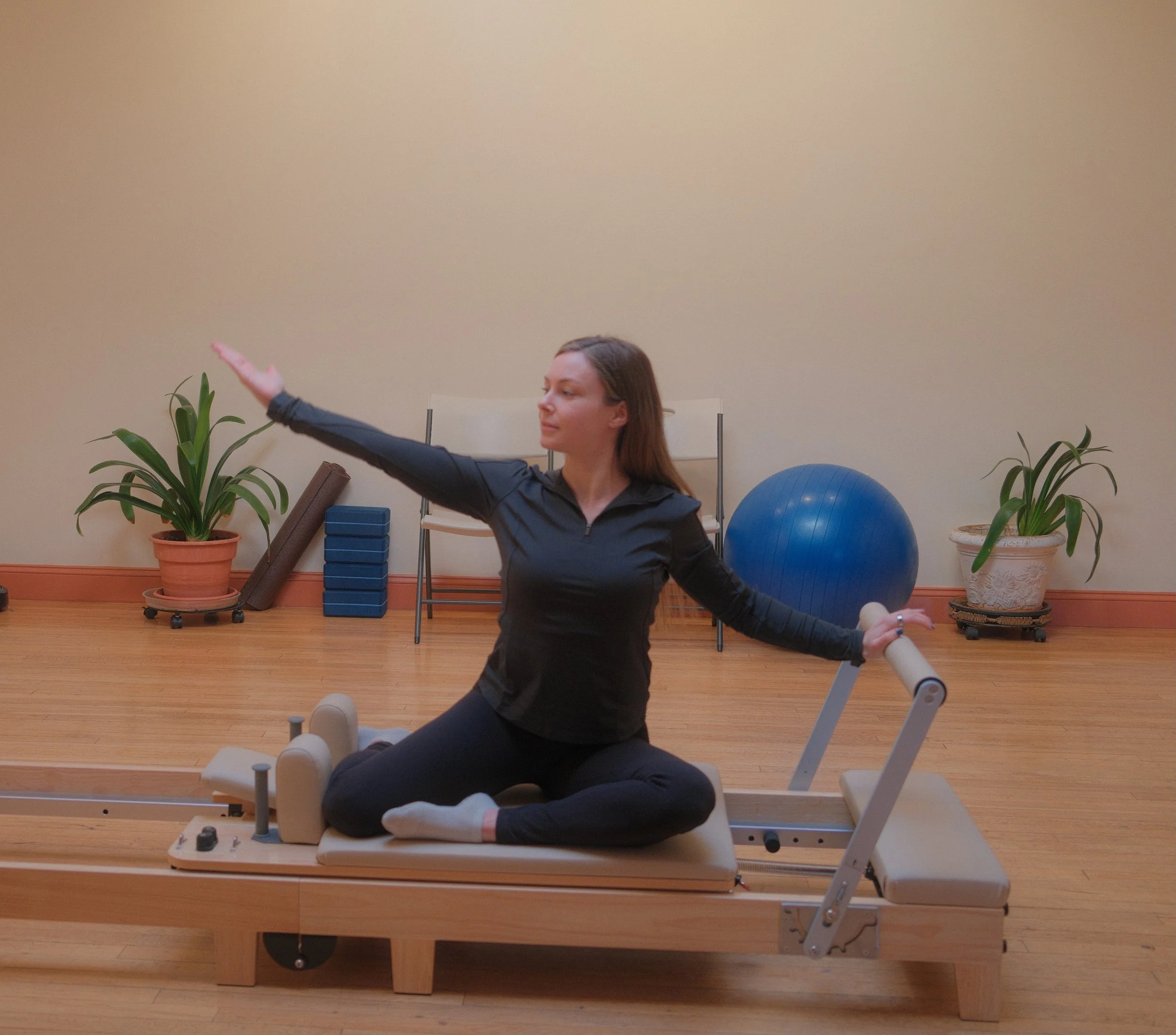 A woman practicing Pilates on a reformer machine in a fitness studio, with exercise equipment, plants, and a large blue exercise ball in the background.