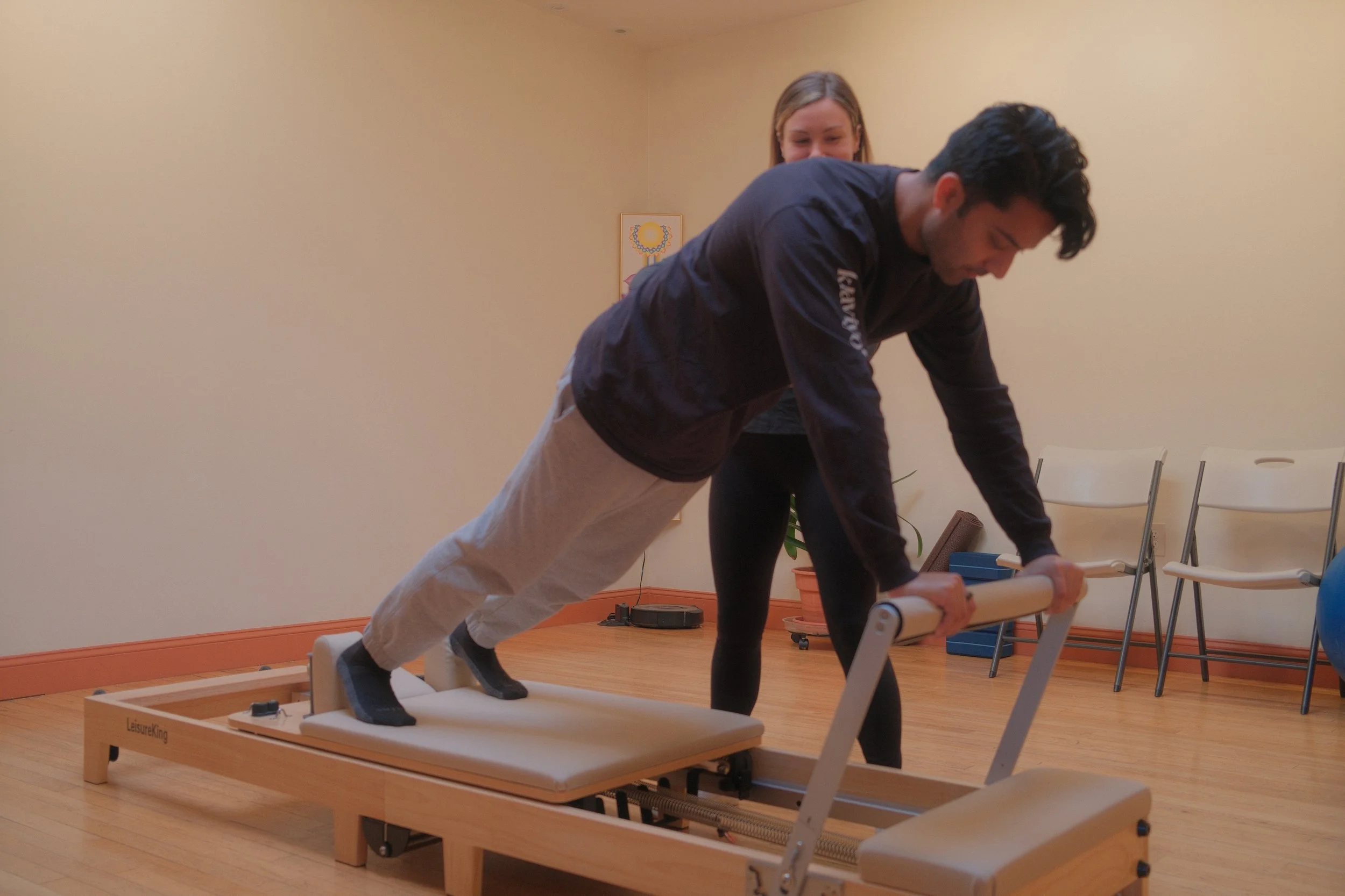 A man doing a plank exercise on a Pilates reformer machine with the assistance of a trainer in a fitness or therapy room.