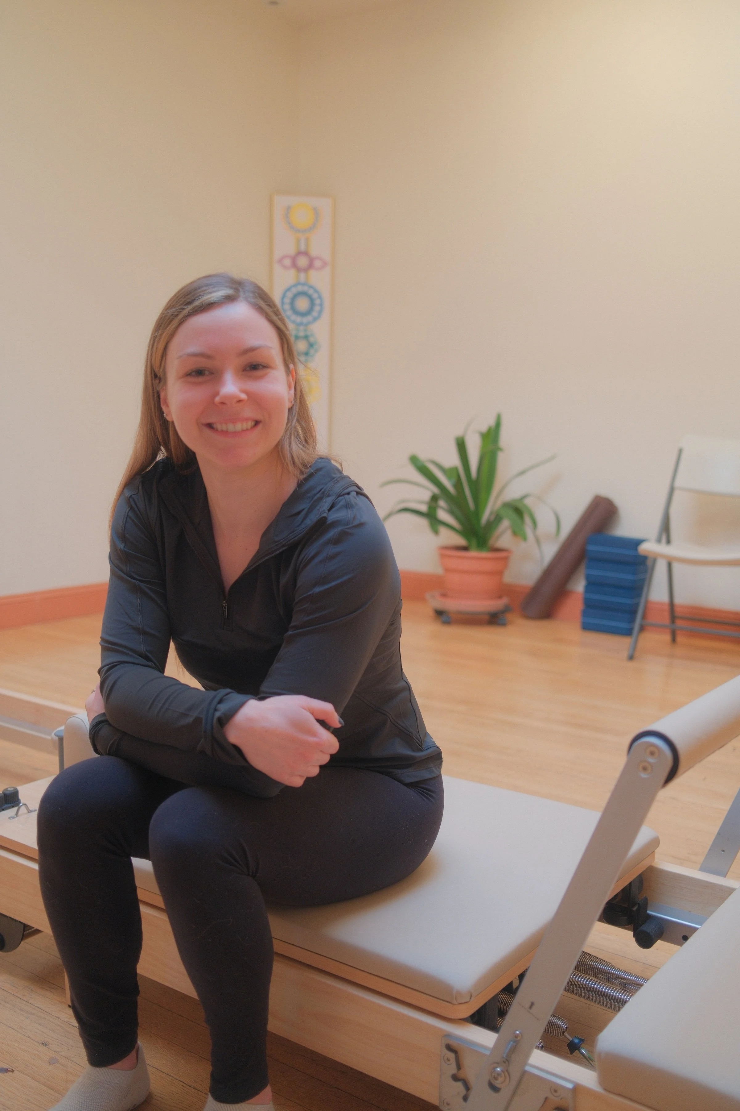 A smiling woman sitting cross-legged on a Pilates reformer machine in a fitness studio, with a potted plant, a folded yoga mat, and a chair in the background.
