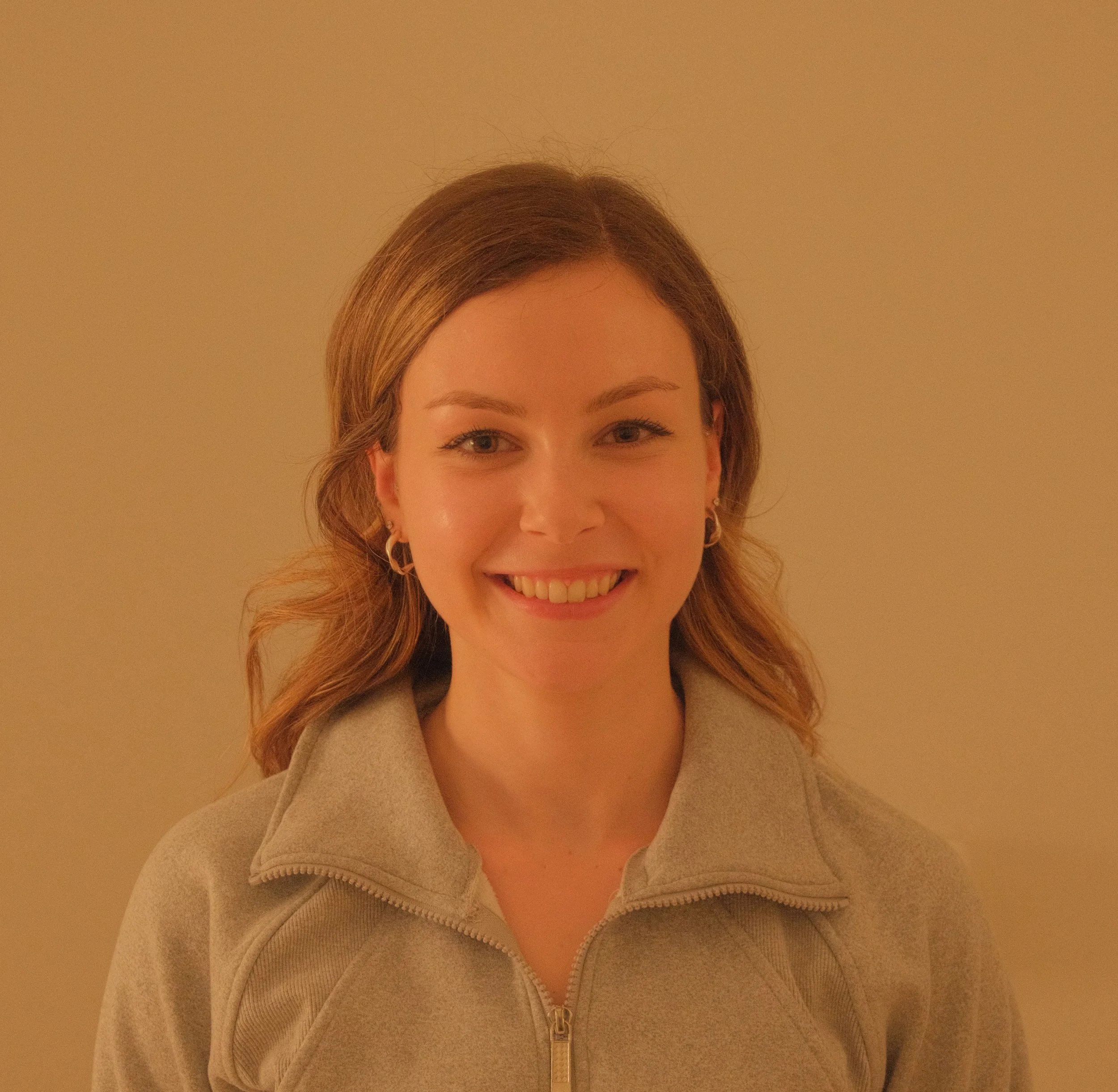 A young woman with light brown hair and earrings, smiling in front of a plain beige wall.